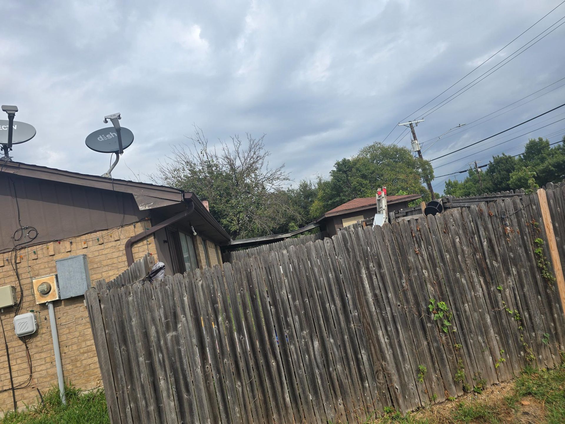 Fenced yard with satellite dishes on a brown roof under a cloudy sky.