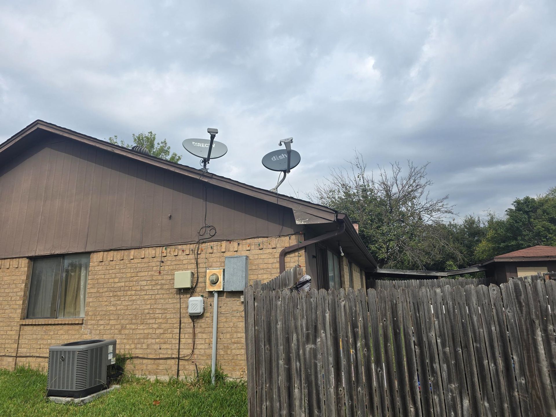 Brown brick building with two satellite dishes on the roof and a wooden fence in the foreground. Overcast sky.