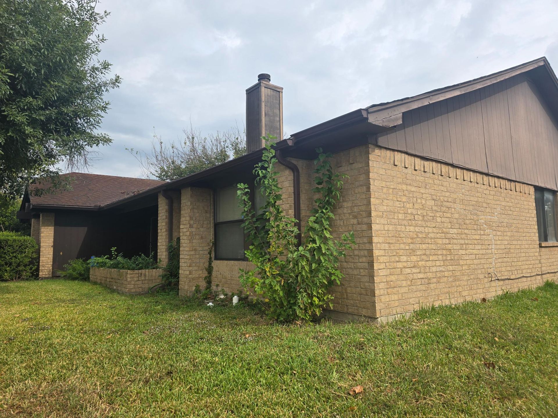 Tan brick house with brown trim and overgrown yard.