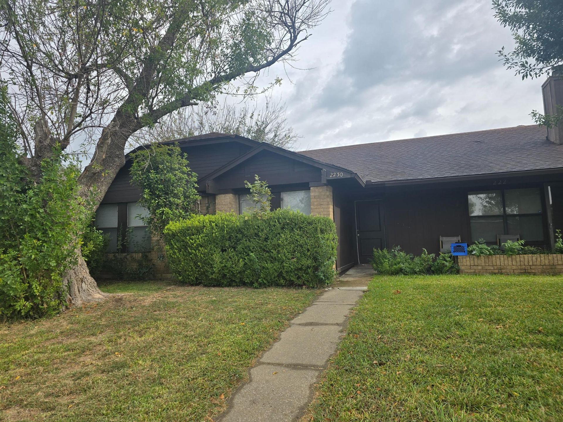 Brown house with brick accents, pathway, and overgrown landscaping under a cloudy sky.