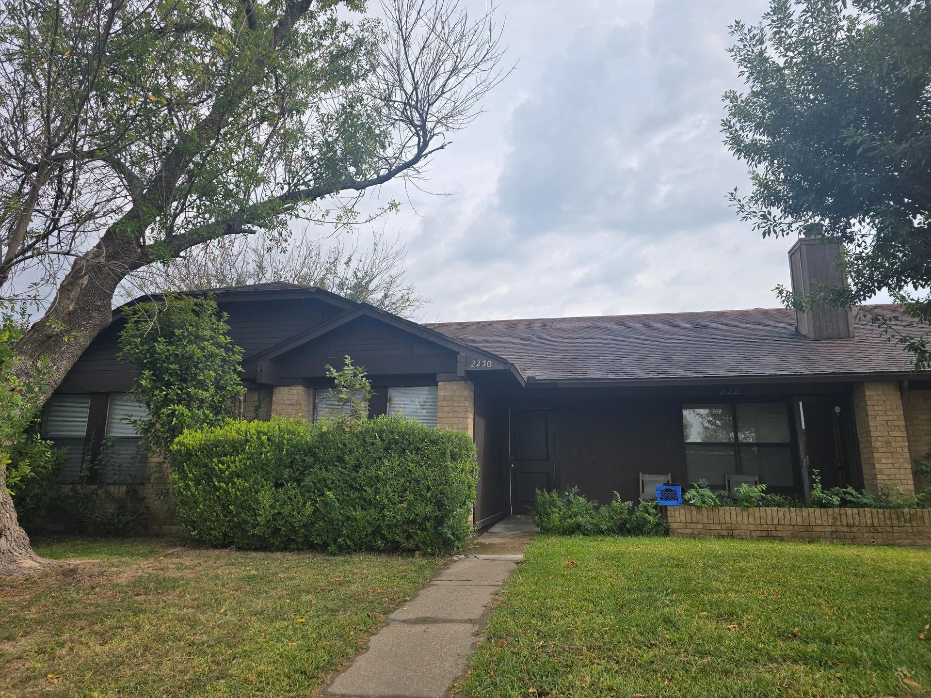 Brown house with brick accents, green lawn, trees, and cloudy sky.