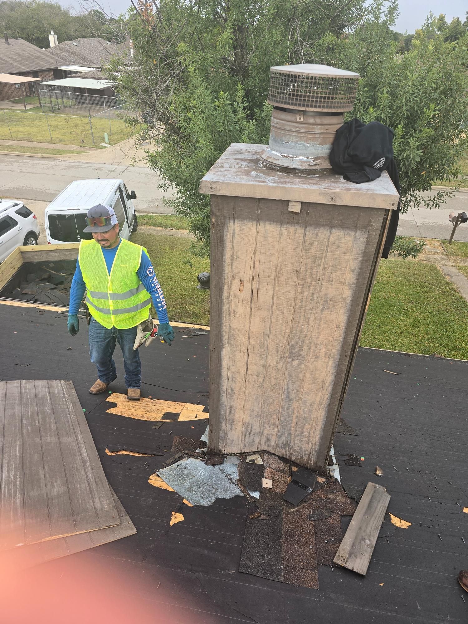 Worker on a roof near a chimney, wearing a safety vest. Damaged roof area with debris. Gray cloudy day.