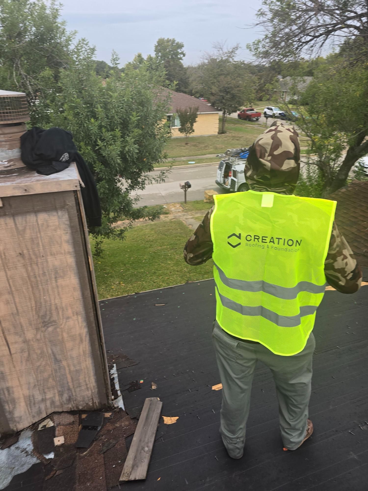 Person on a rooftop wearing a high-visibility vest inspects roofing, suburban setting.