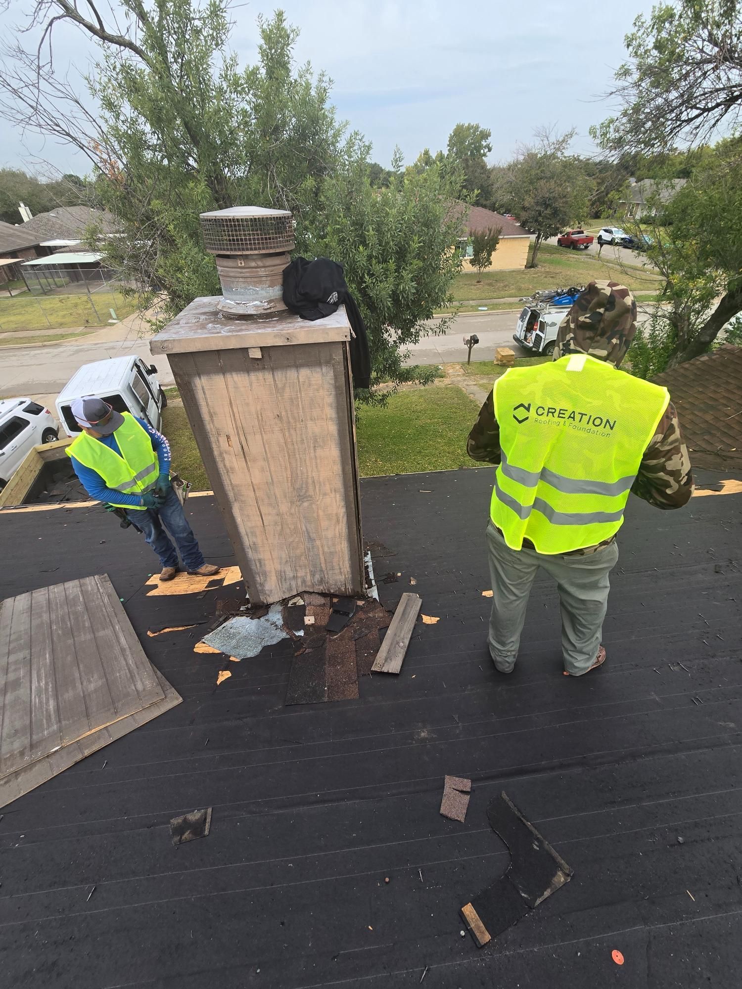 Two workers on a roof next to a chimney. One in a yellow vest is looking down.