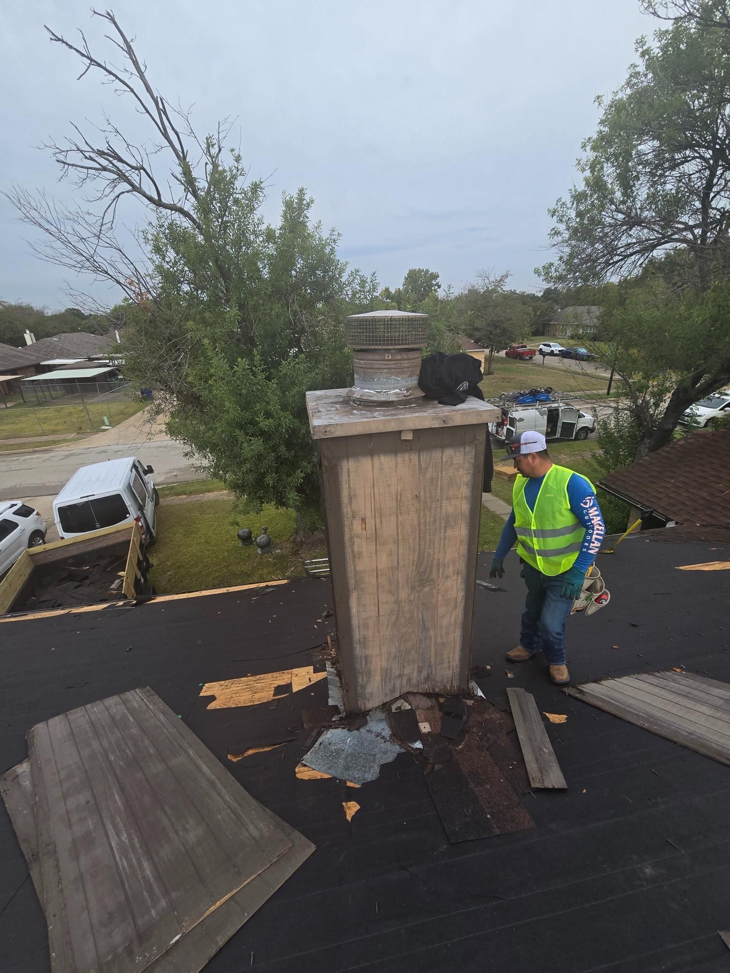 Person on a roof with a chimney, wearing a safety vest. Torn roofing materials are visible. Cloudy sky.
