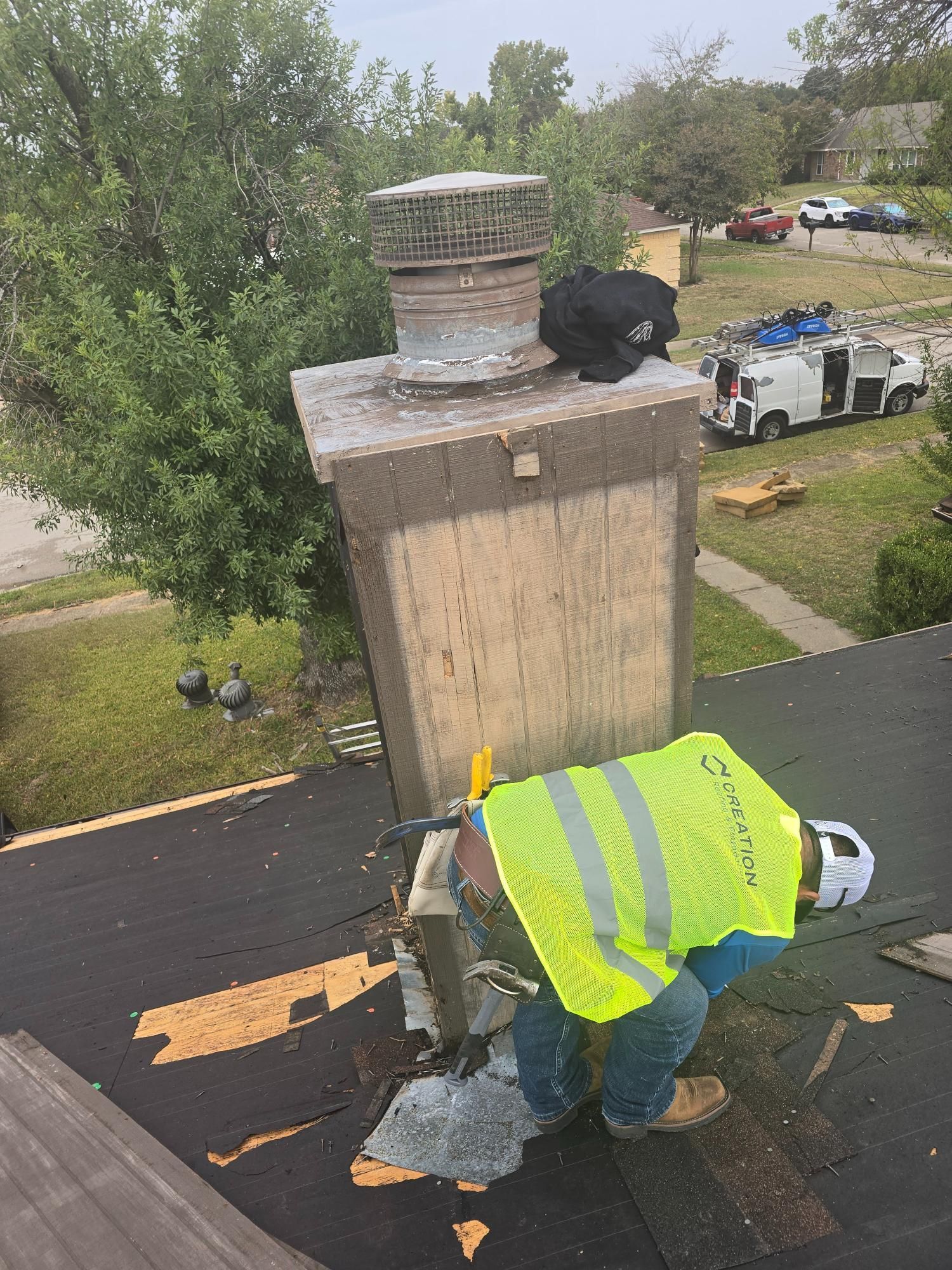 Roofer in safety vest using a saw to cut around a chimney on a dark roof.
