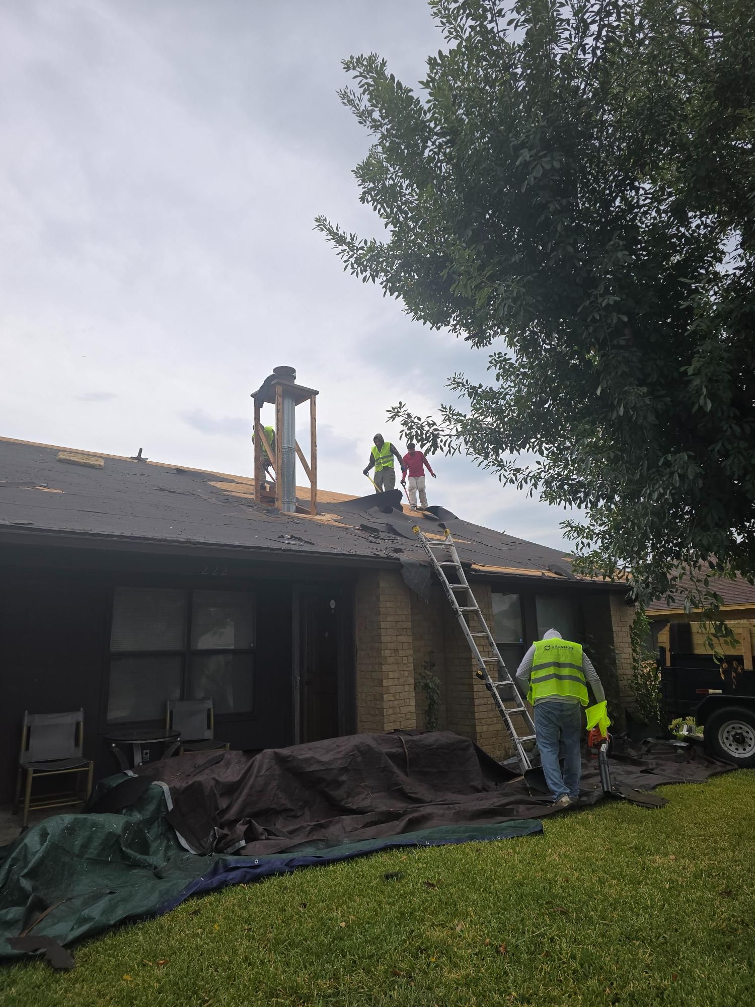 Roofers on a house with exposed roof. One climbs ladder, two on the roof near chimney. Green tarp covers the grass.