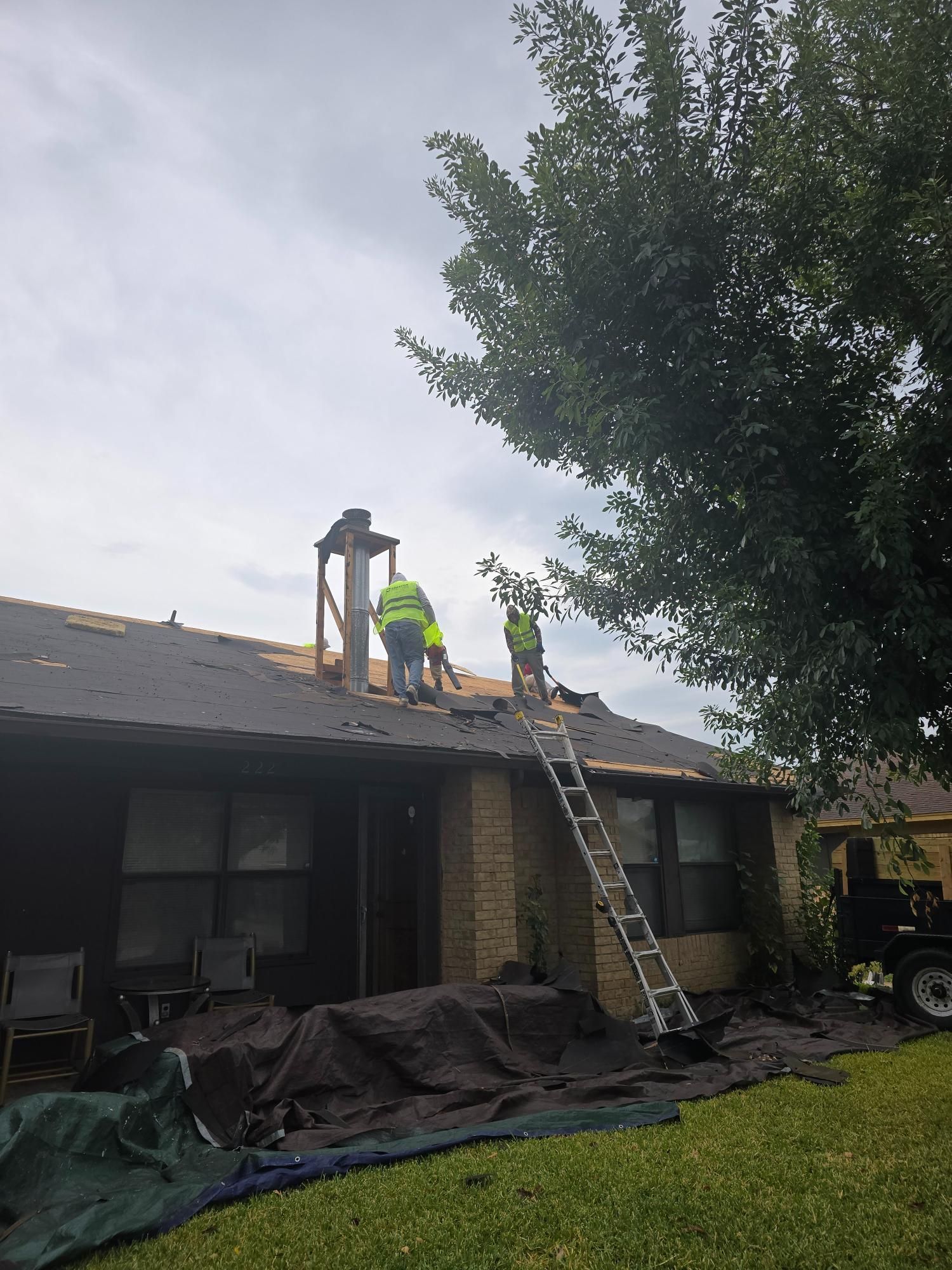 Roofing crew replacing shingles on a house, working near a chimney and ladder on a cloudy day.