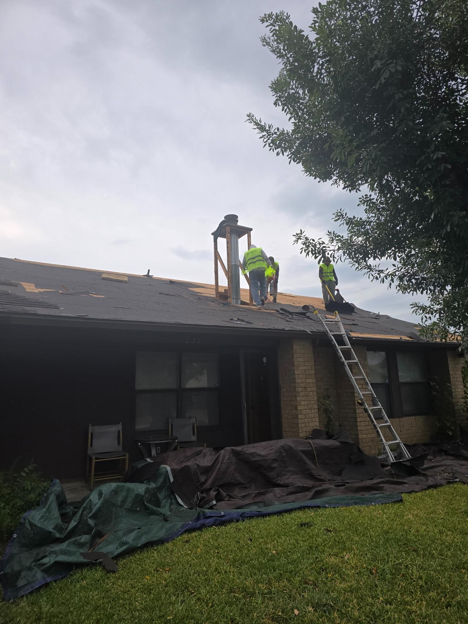 Roofers on a house working near a chimney, ladder leaned against the roof, tarp in the foreground.
