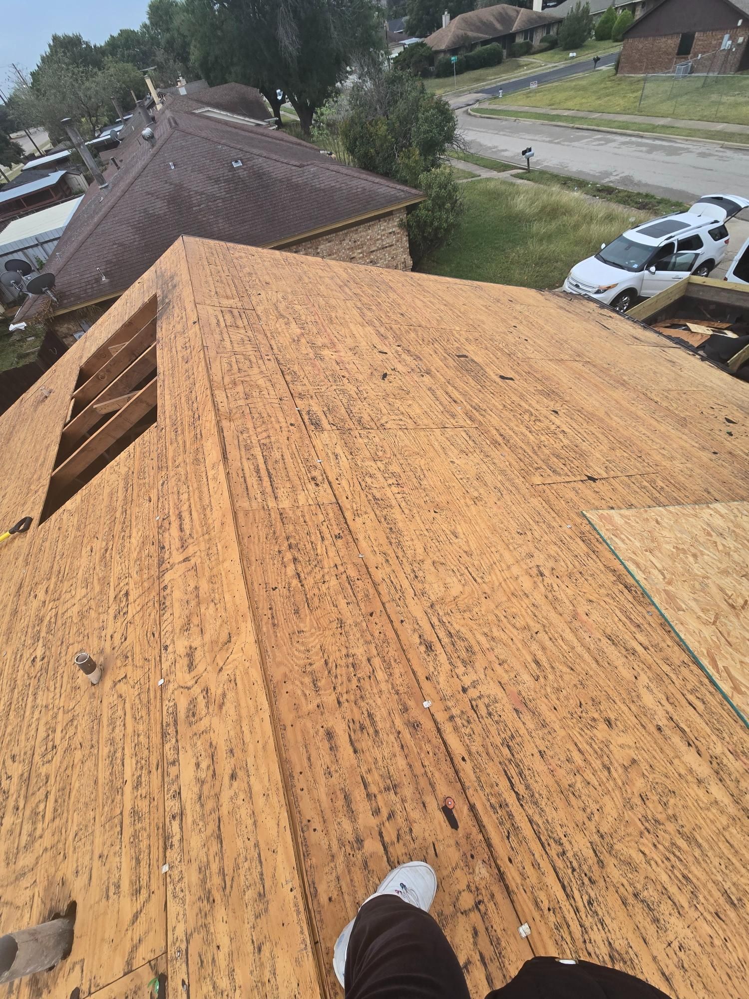 Rooftop with exposed wooden sheathing, white sneakers visible. Houses and street in background.
