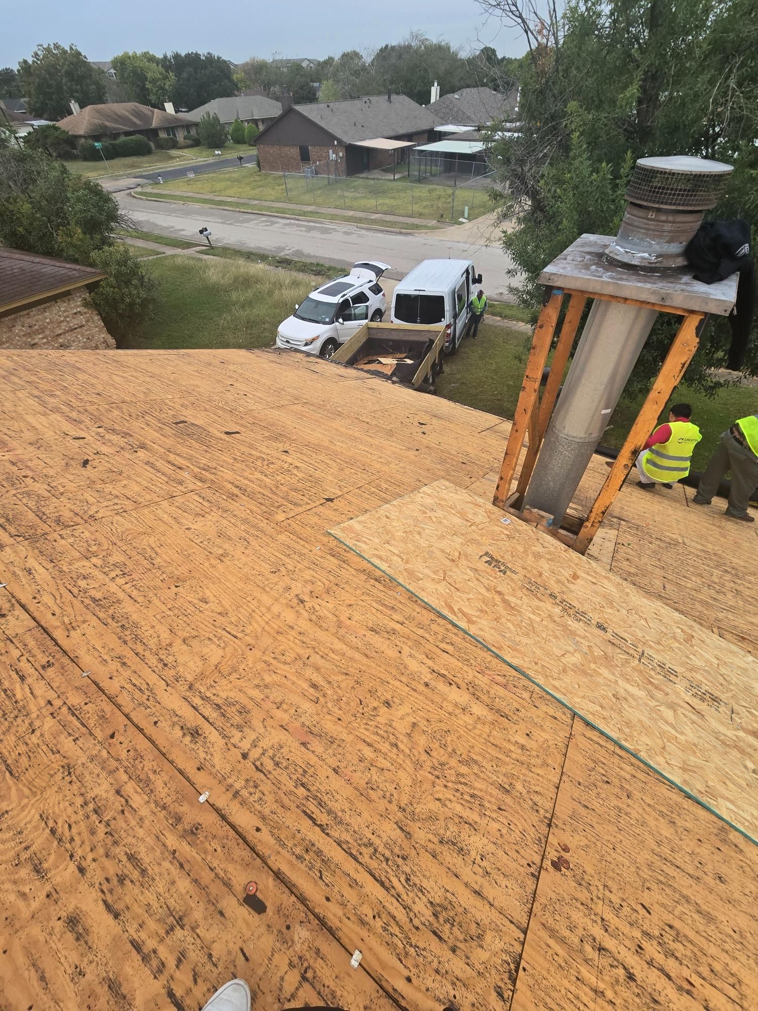 Roof being repaired, partially covered with new wood. Workers, vehicles, houses in the background.