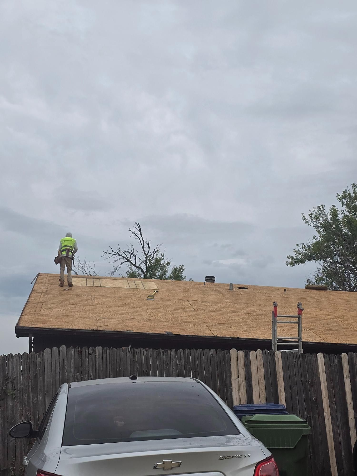 Roofer on a house roof under a cloudy sky, removing old shingles. A car and fence are in the foreground.