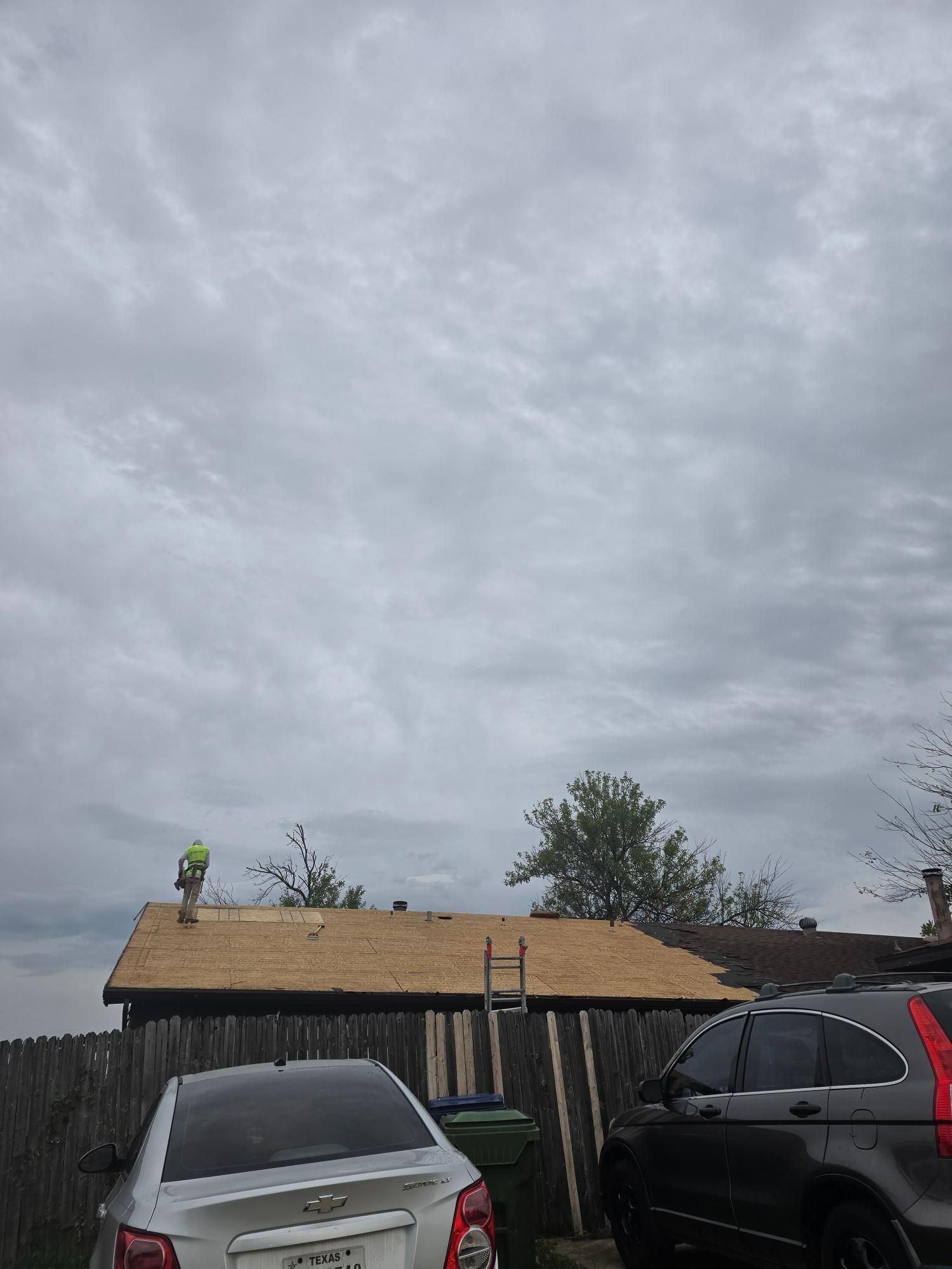 Person on a roof, working. A car and fence are in the foreground, cloudy sky above.