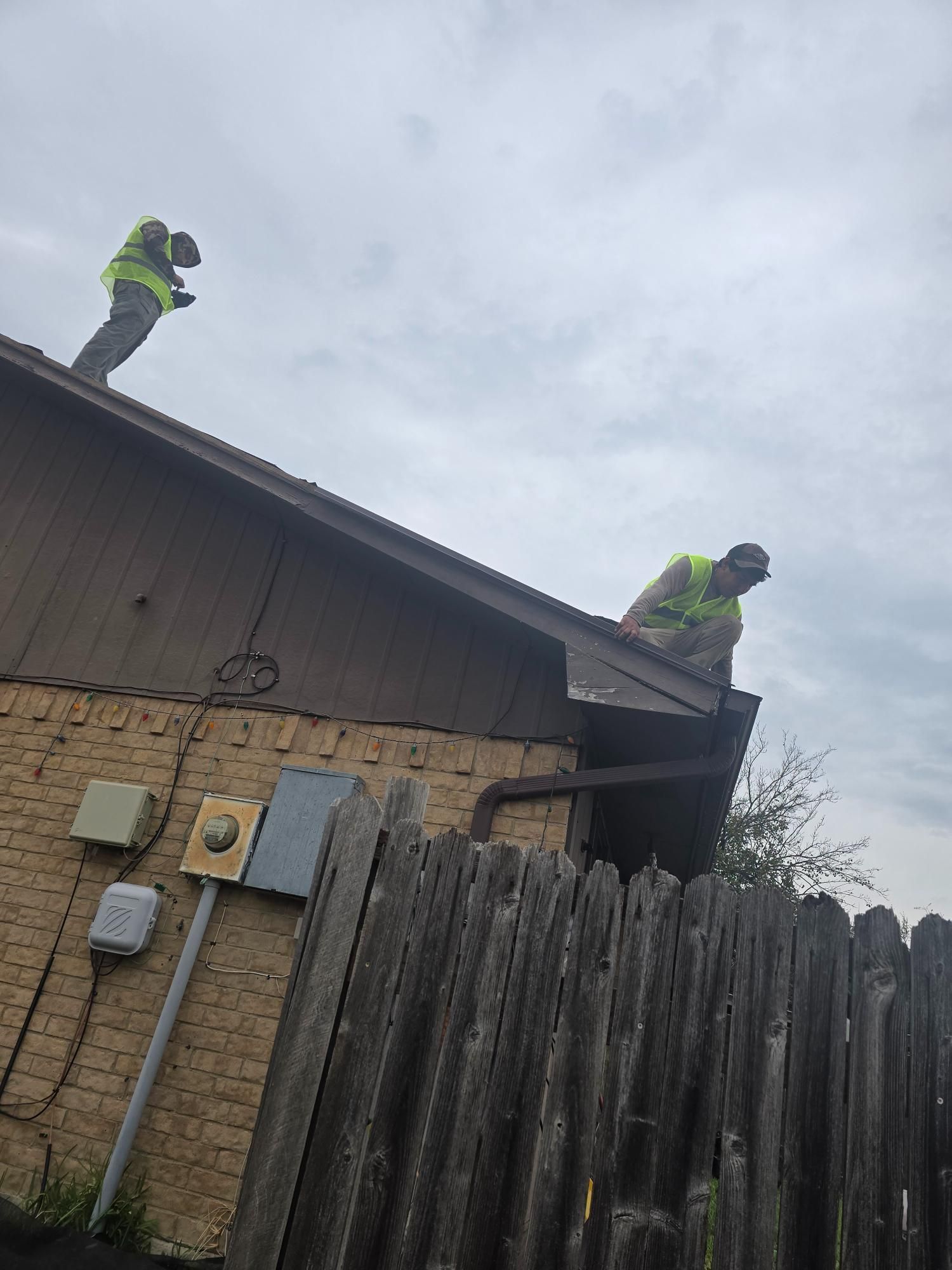 Two people in safety vests on a brown roof near a wooden fence. Overcast sky.