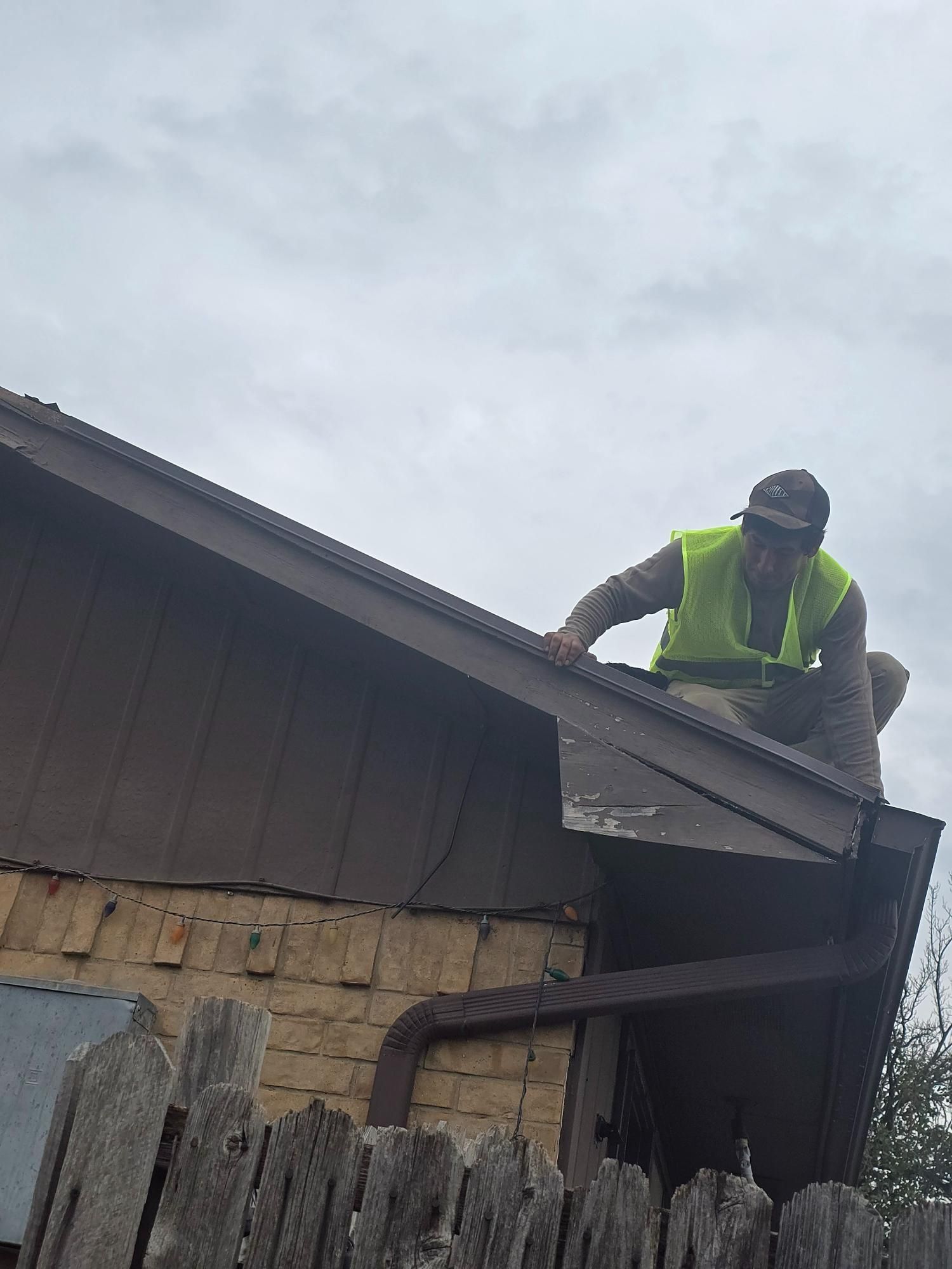 Person wearing a safety vest on a brown roof, working on its edge against a cloudy sky.