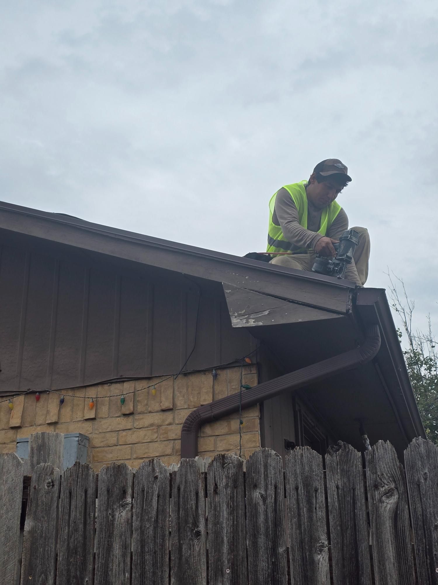 Person on a roof wearing a safety vest. Brown house with a gutter and a wooden fence below. Cloudy sky.