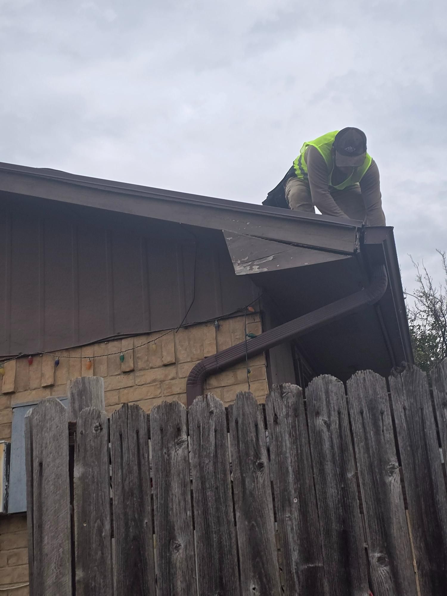 Person on a roof, in a yellow vest, working on a gutter above a wooden fence. Overcast sky.