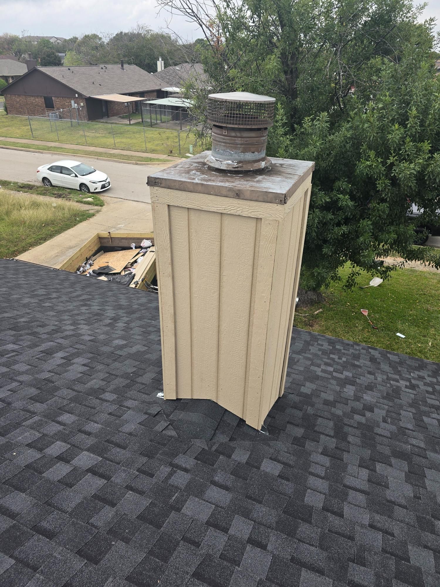 Chimney on a dark shingle roof with a metal cap, light tan siding, and a suburban background.