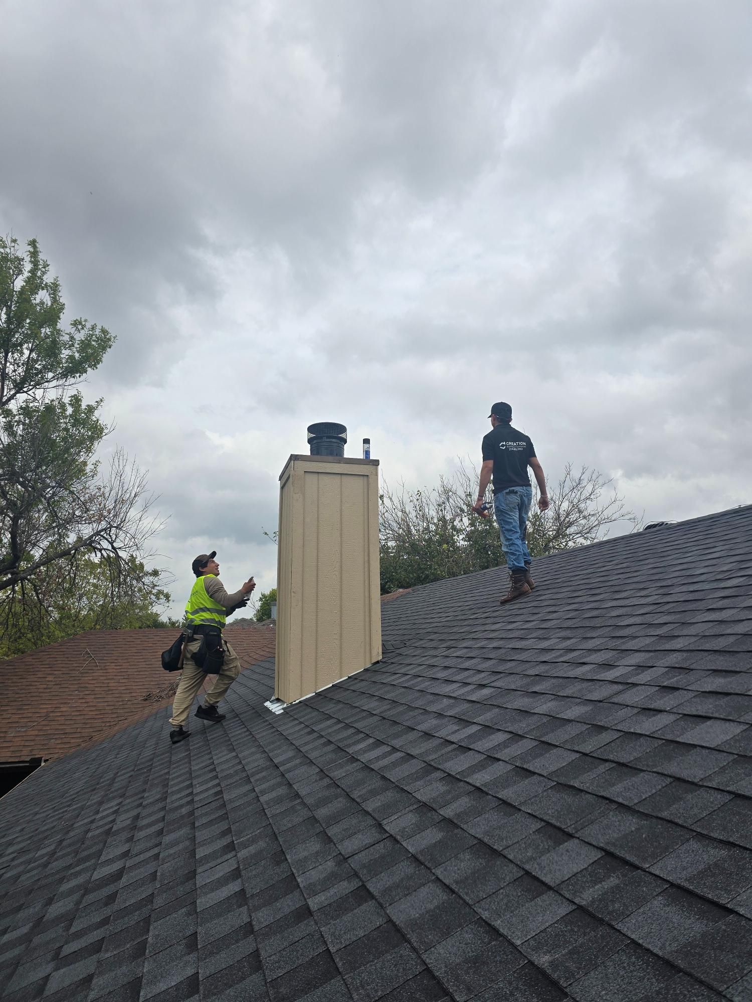 Two roofers on a dark shingle roof near a chimney under a cloudy sky. One points, the other walks.