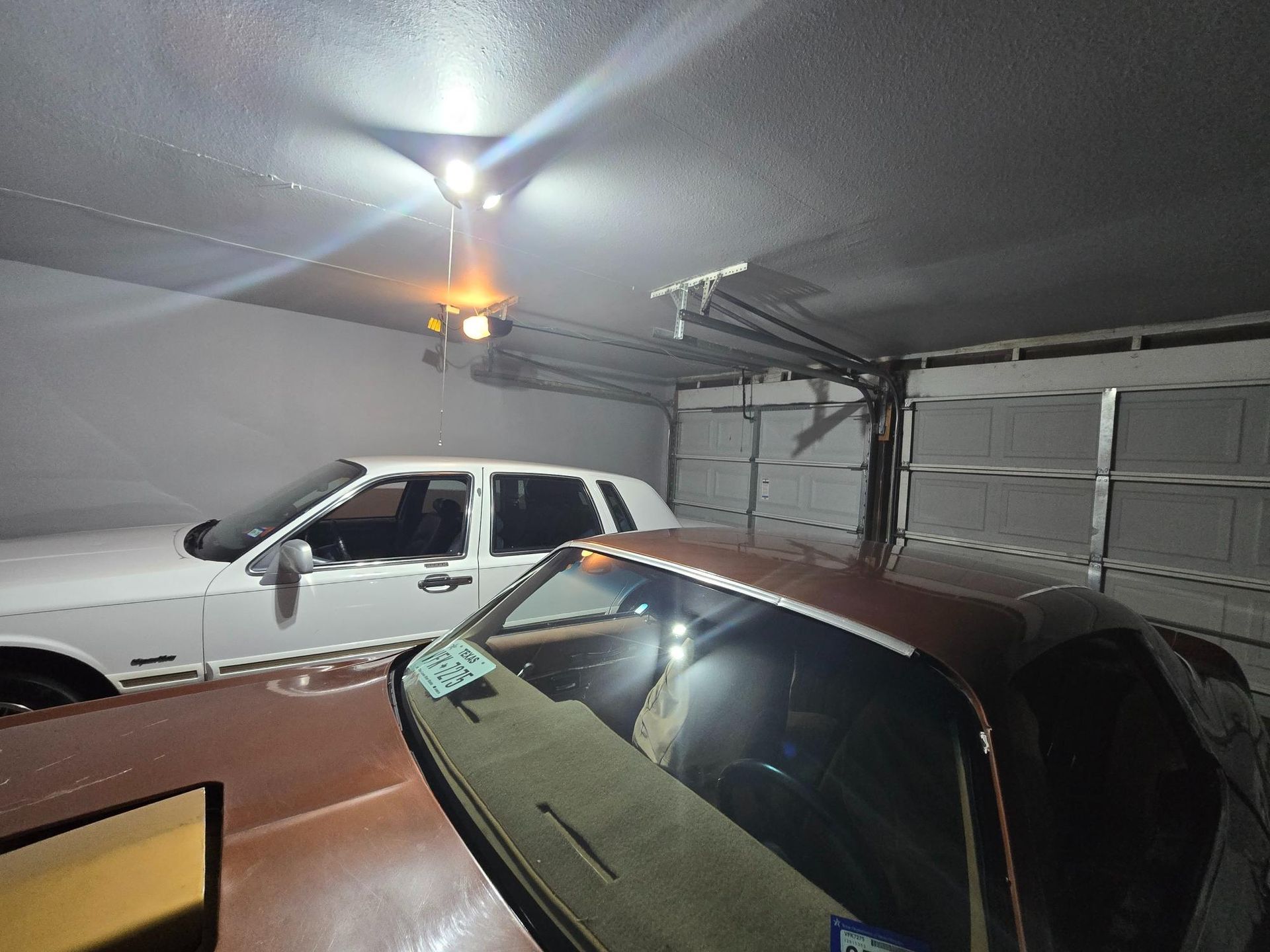 Two cars parked in a garage with the garage door open. Overhead lights illuminate the space.
