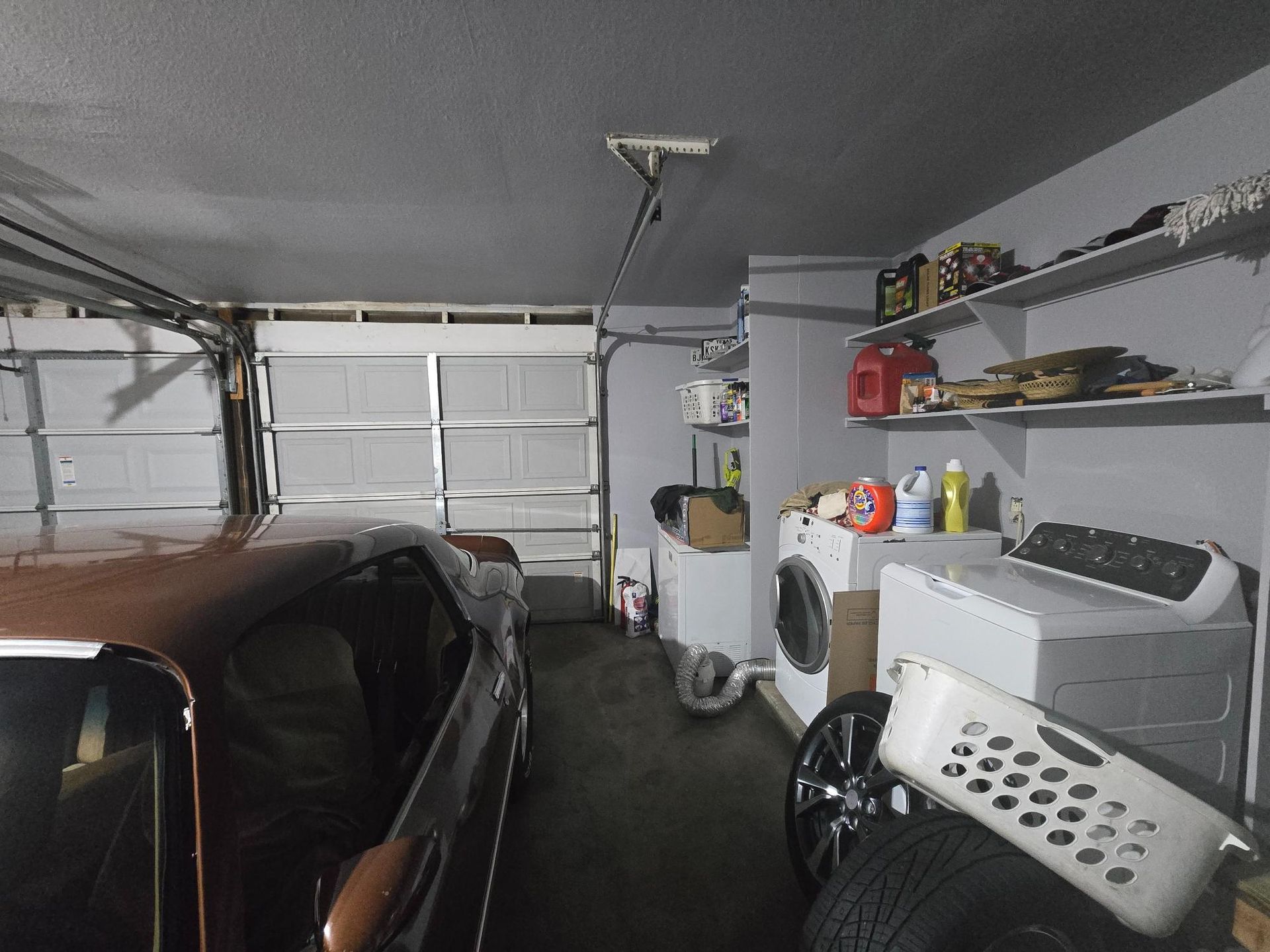 A car parked in a garage with a washing machine, shelves, and a garage door open to the outside.