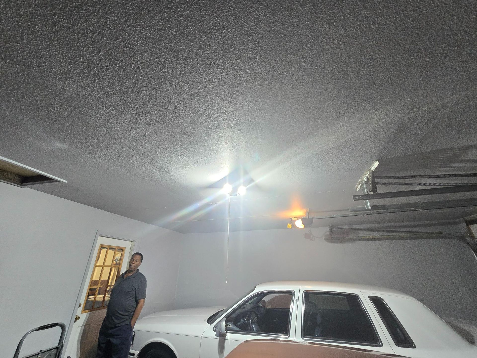 Man standing in a garage with a white car. The ceiling has a textured pattern.
