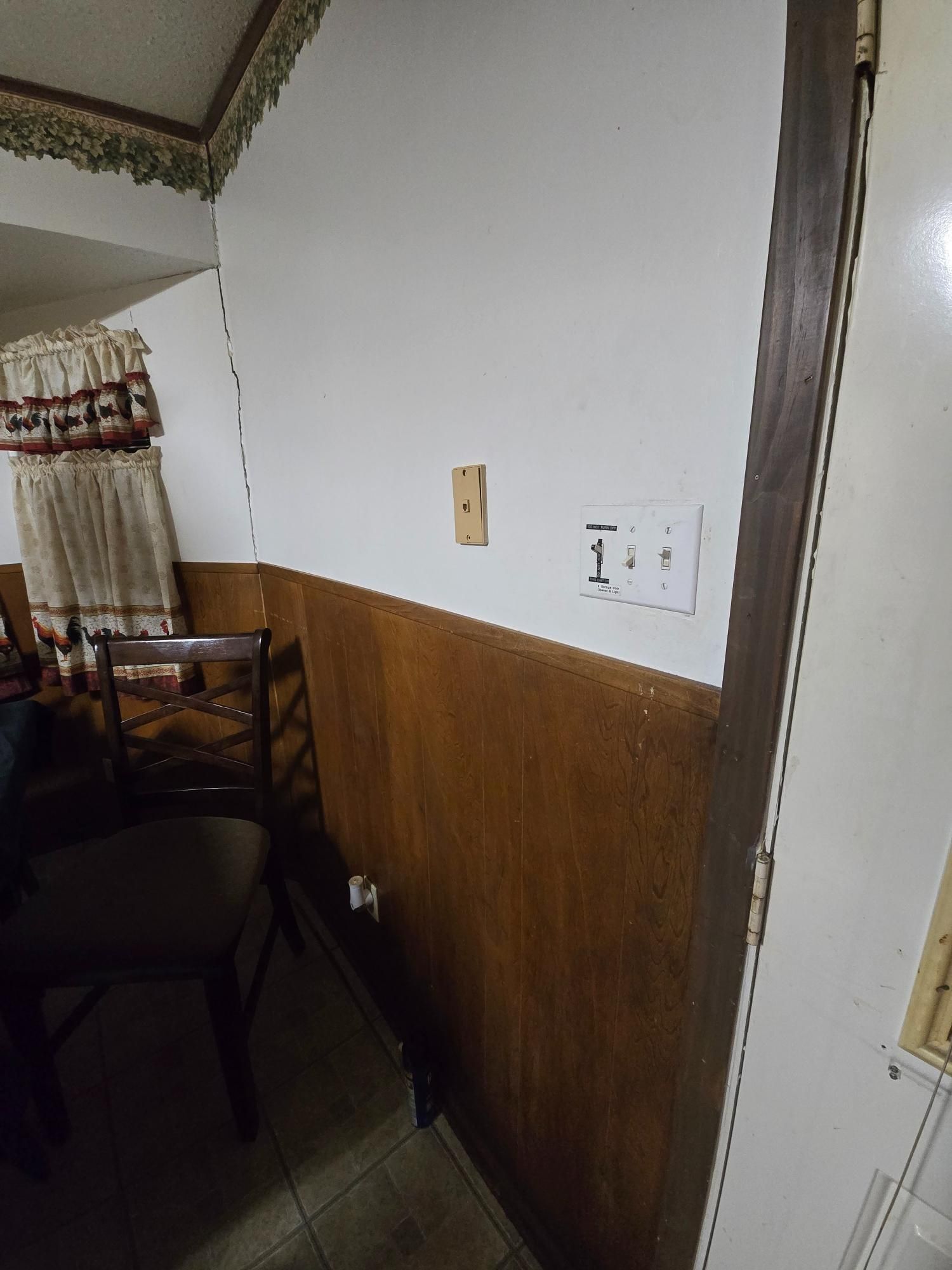Wall with wood paneling, outlet, light switch, and doorway. Dining room setting with chair and curtains.