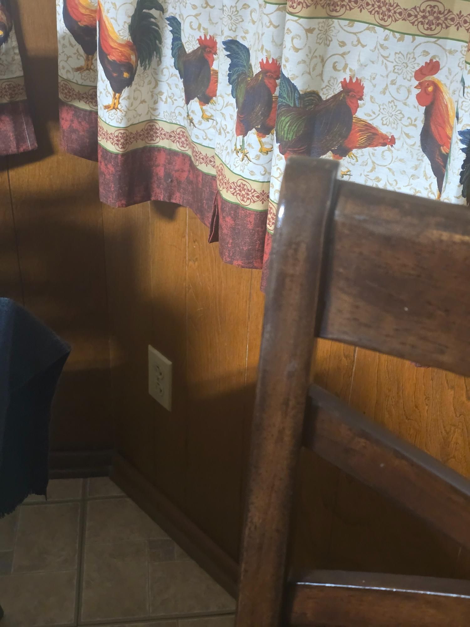 Wooden chair in front of a wall with rooster-themed curtains.  The walls are paneled and tile is on the floor.