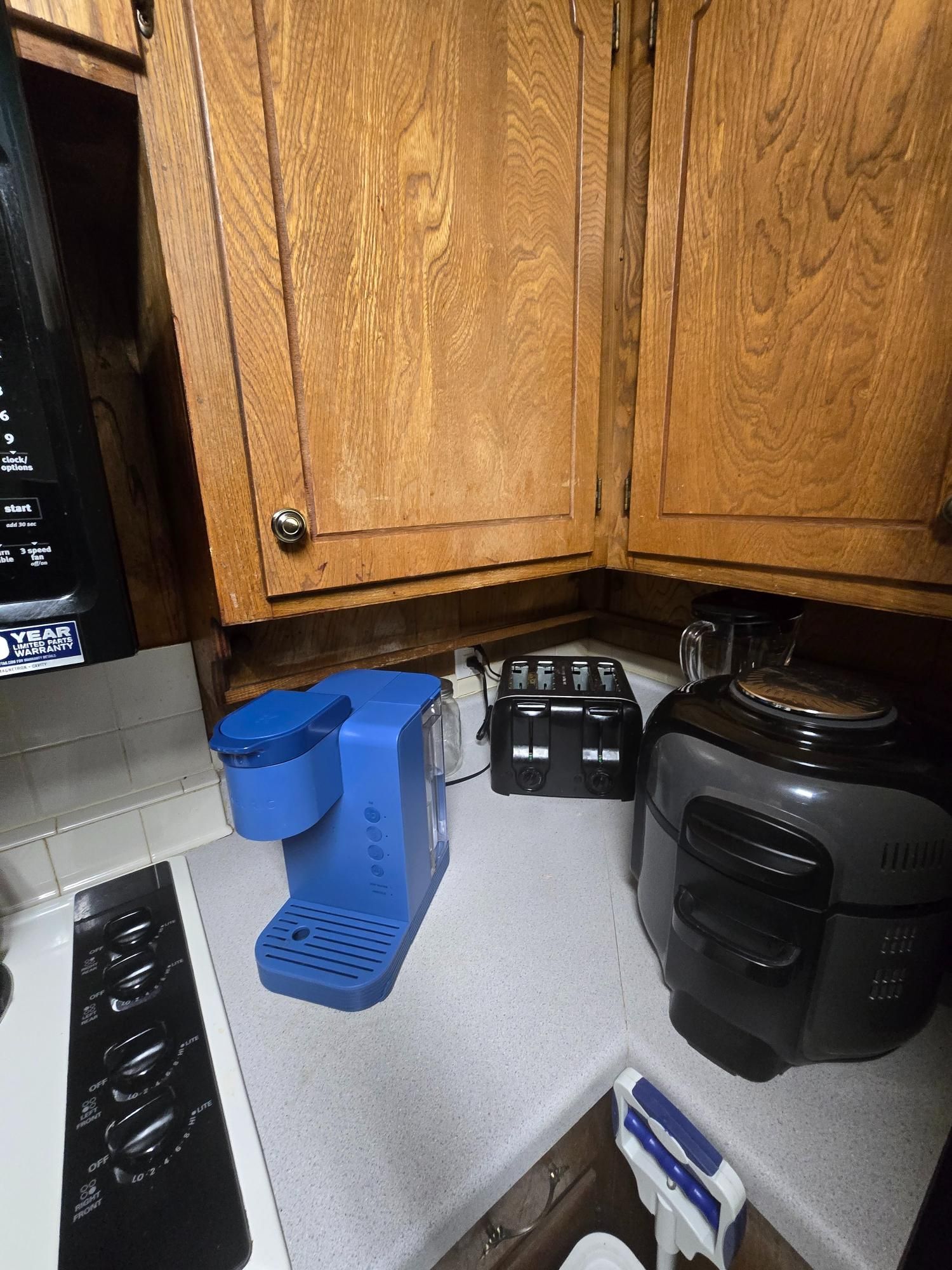 A kitchen countertop with a blue Keurig, black toaster, and gray appliance under wooden cabinets.