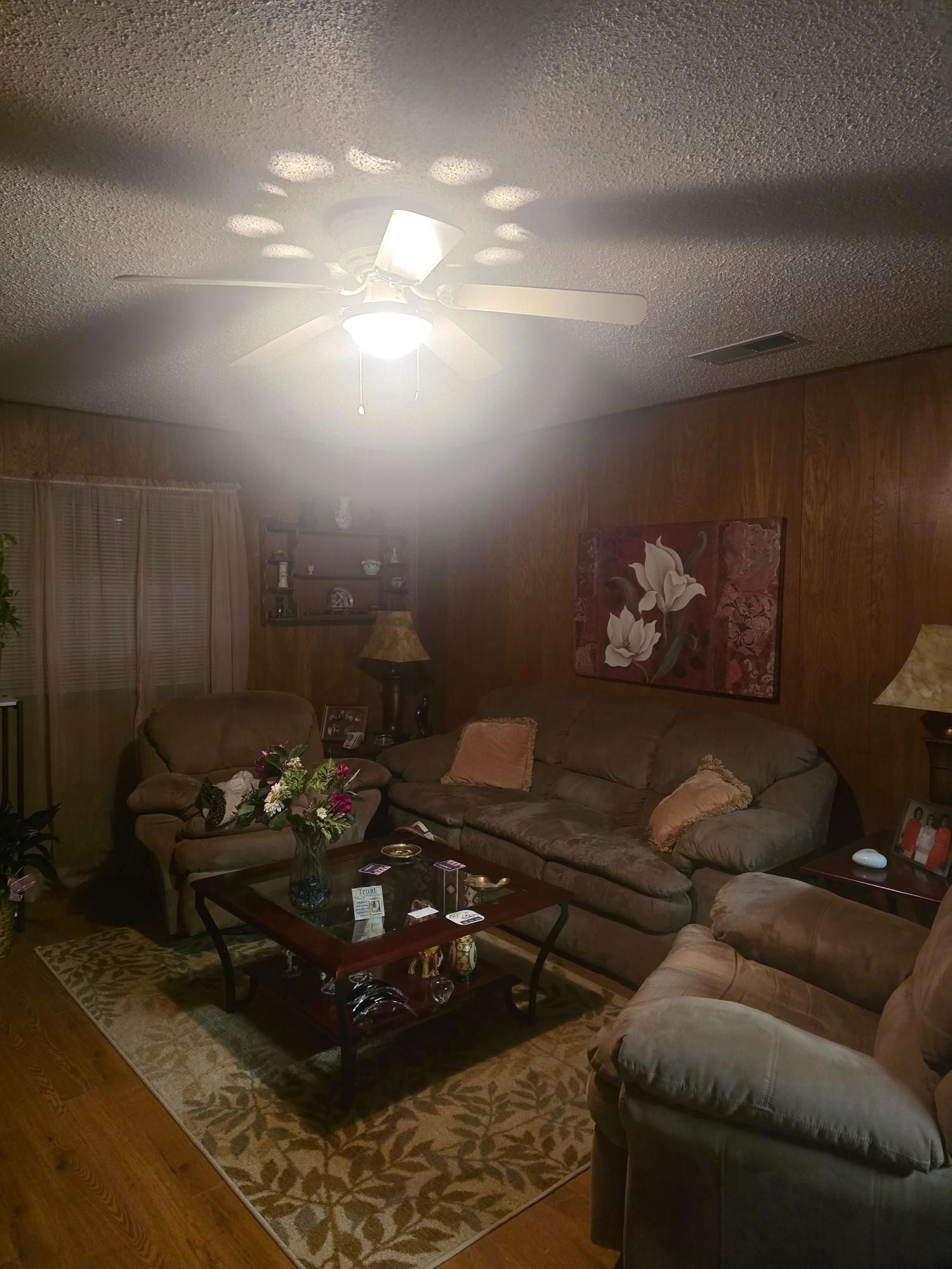 Living room with brown paneling, sofas, coffee table, and area rug. White ceiling fan and flower art.