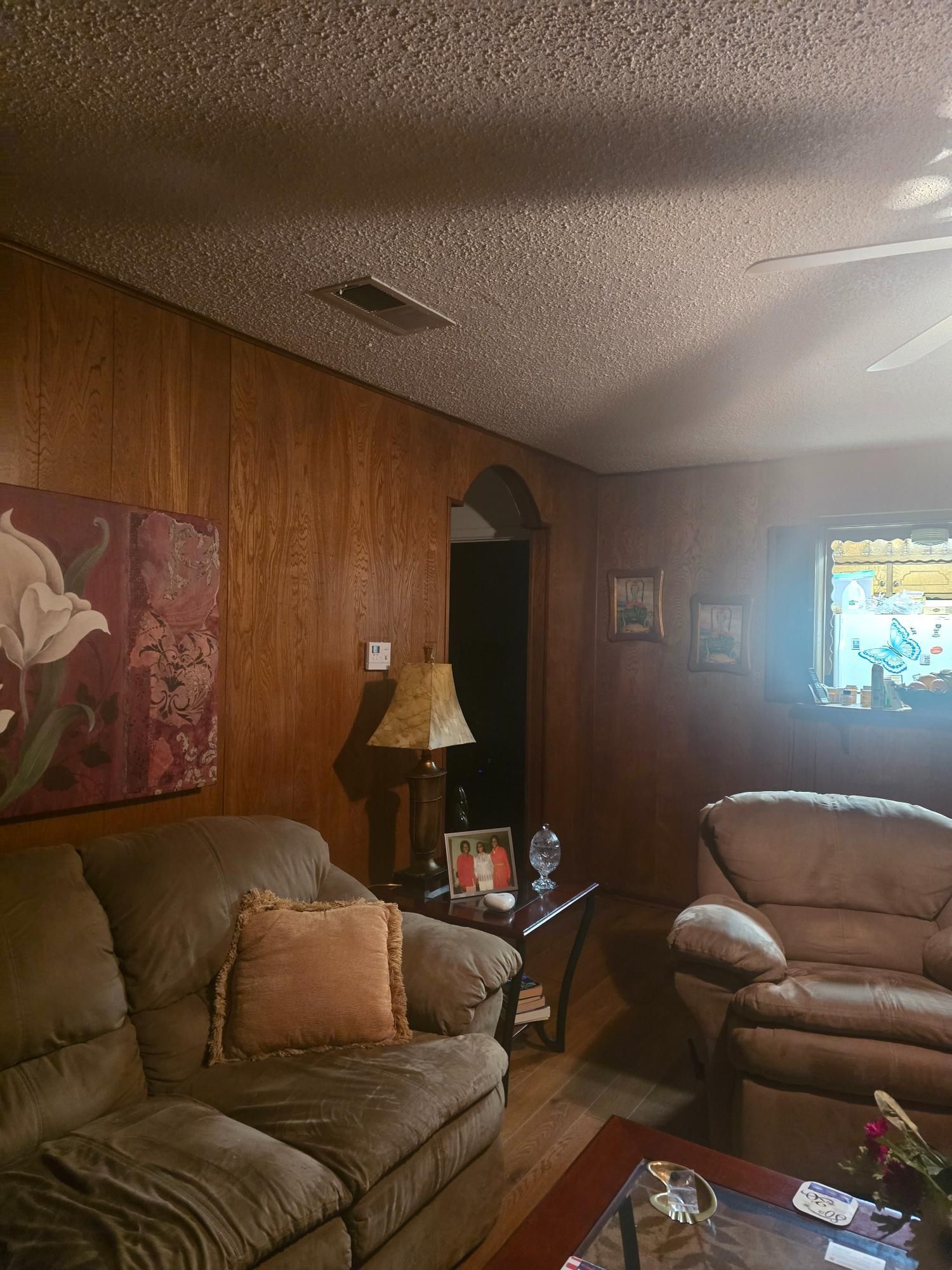 Living room with wood paneling, brown sofa and chair, floral artwork, and a table with a lamp.