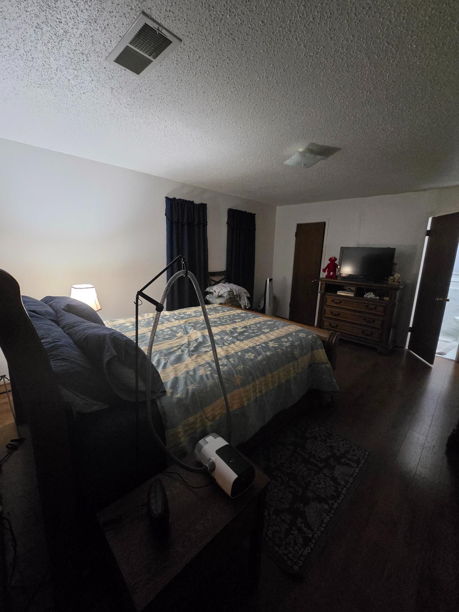 Bedroom with bed, dresser, TV, and open door to the outside. Dark wood floors and white walls.
