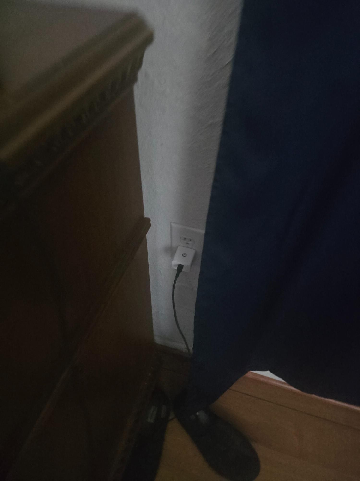 An electrical outlet with a phone charger plugged in, next to a nightstand and curtain.