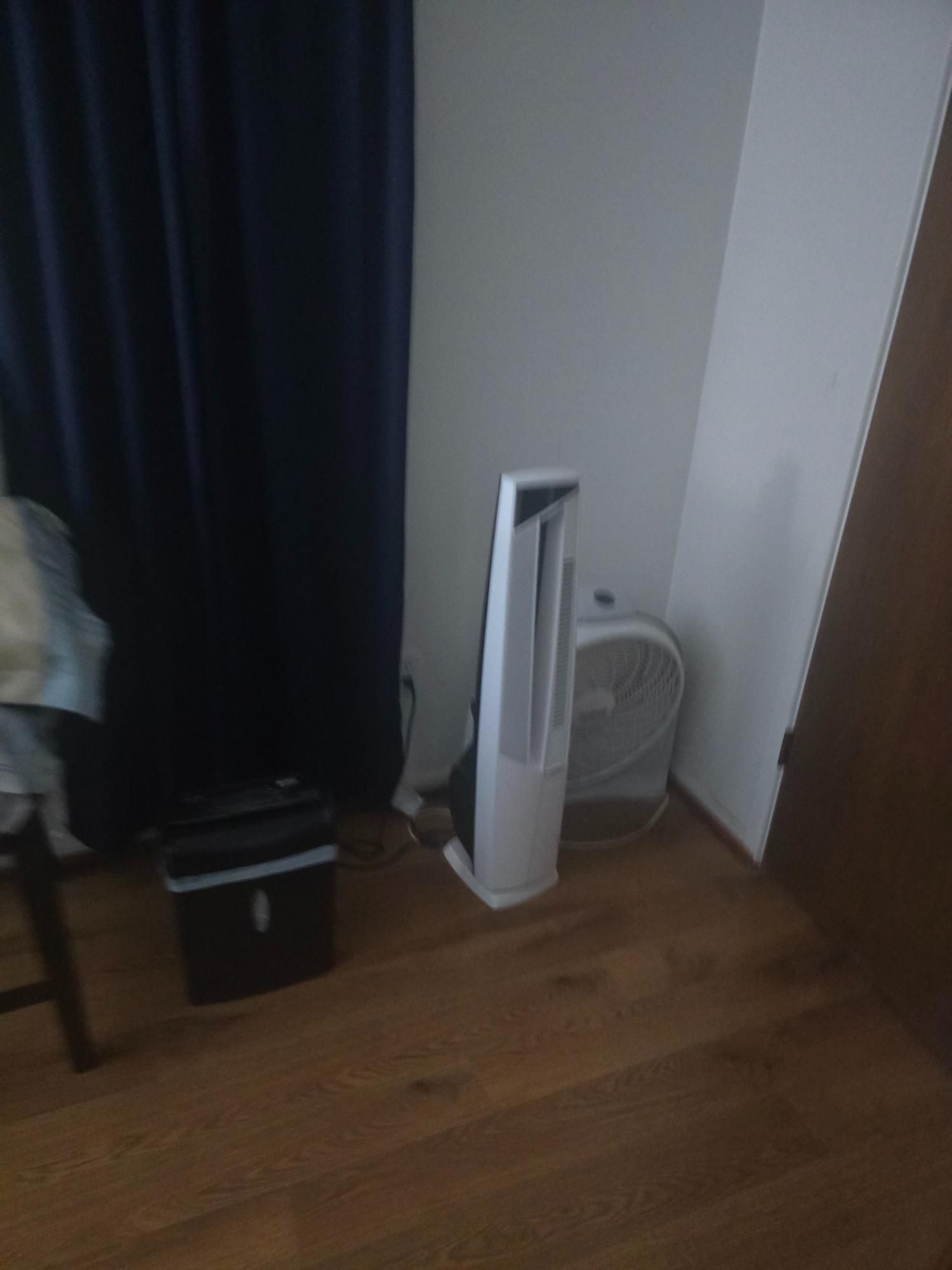 A room corner with a white tower fan, a box fan, a black shredder, and a blue curtain.