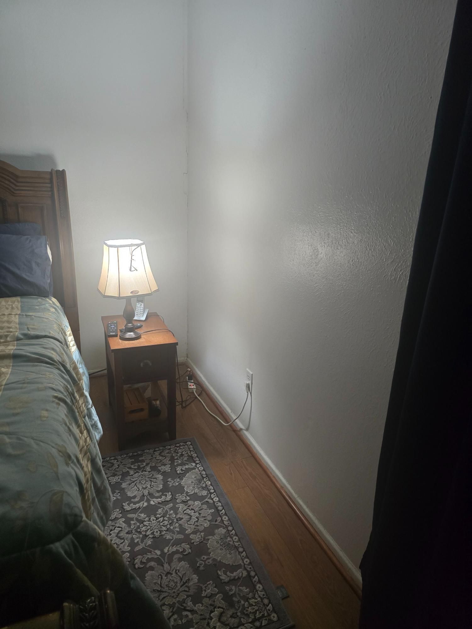 Bedroom corner with a wooden nightstand, lamp, and rug. Bed is on the left; a dark curtain is on the right.