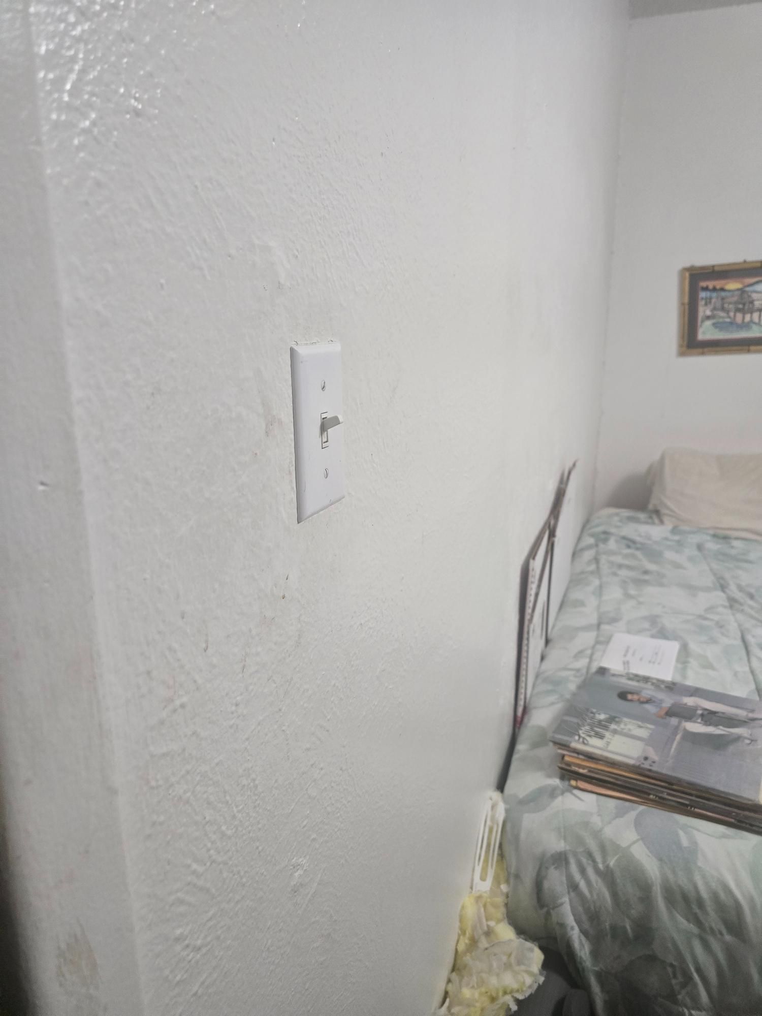 White wall with a light switch, next to a bed with a floral bedspread.