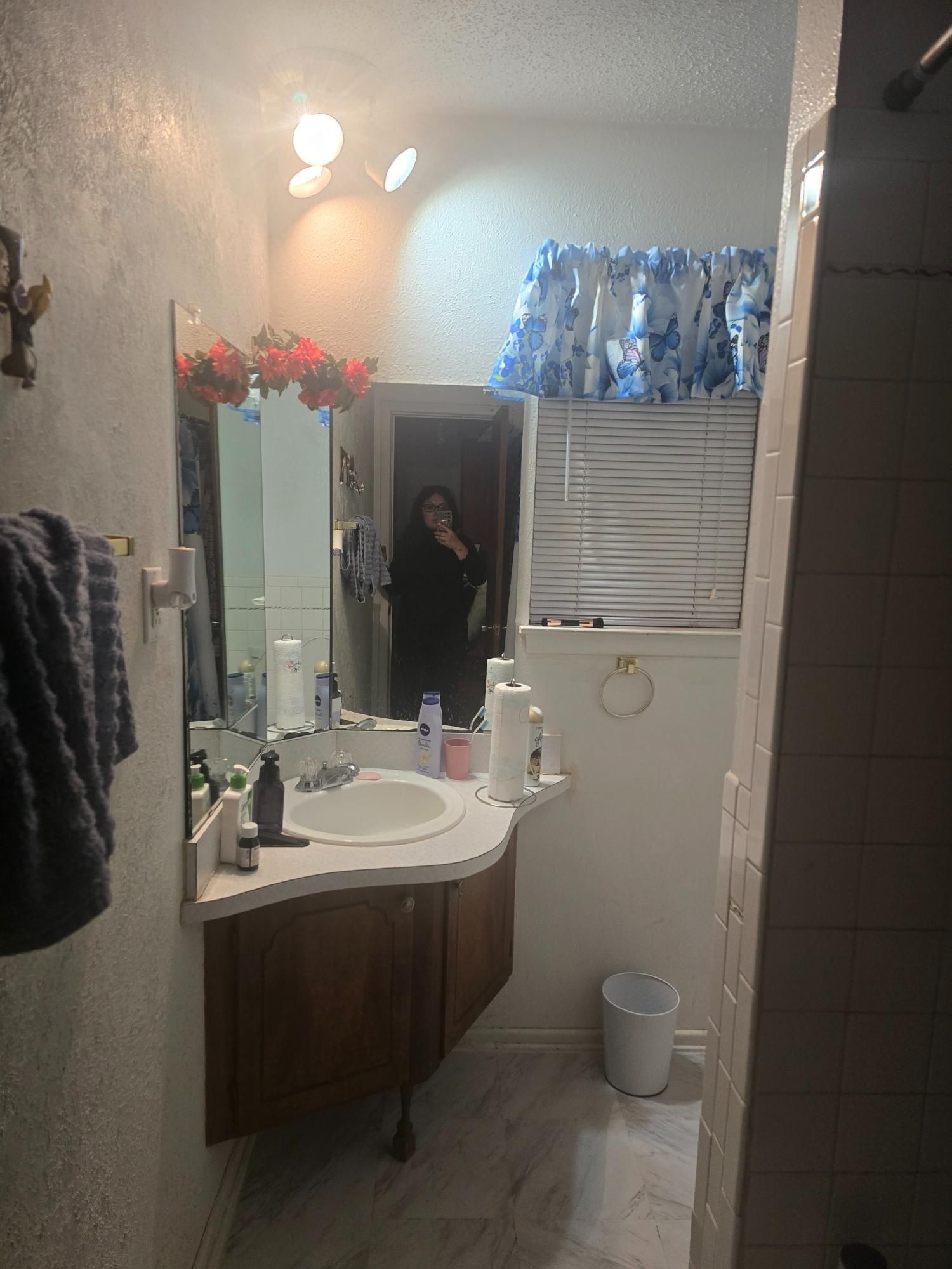 Bathroom interior with sink, mirror, and a person in the mirror. White walls, wooden cabinet, and blue floral curtains.