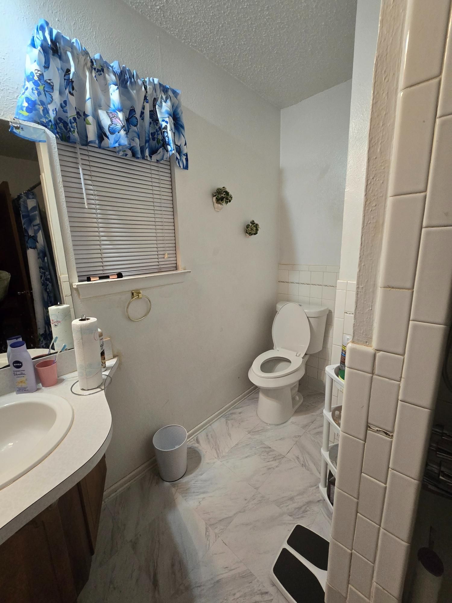 Bathroom interior with toilet, sink, and shower. White walls, floral curtain, and patterned floor.