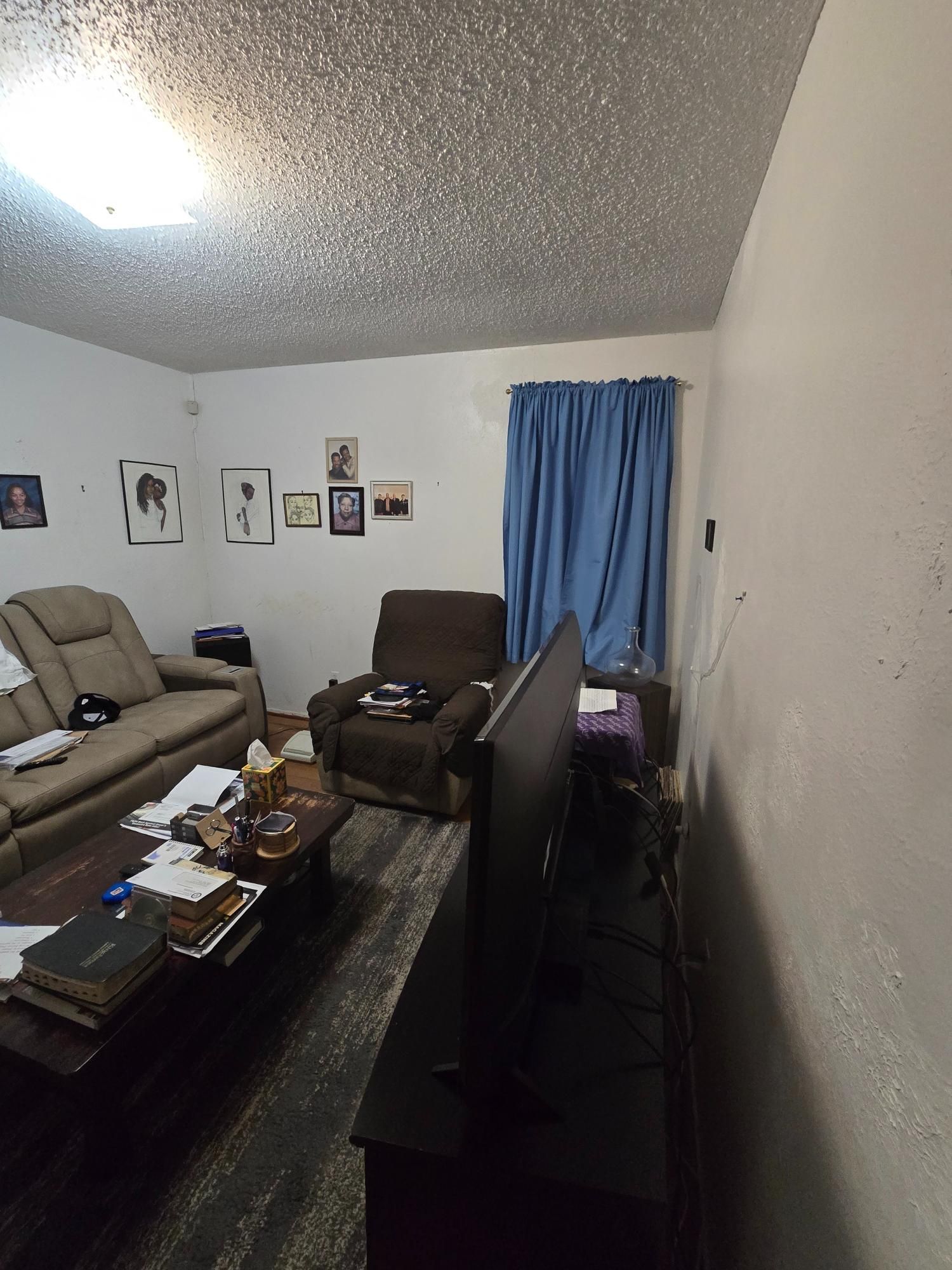 Living room with couch, recliner, coffee table, TV, and blue curtains. White walls and textured ceiling.