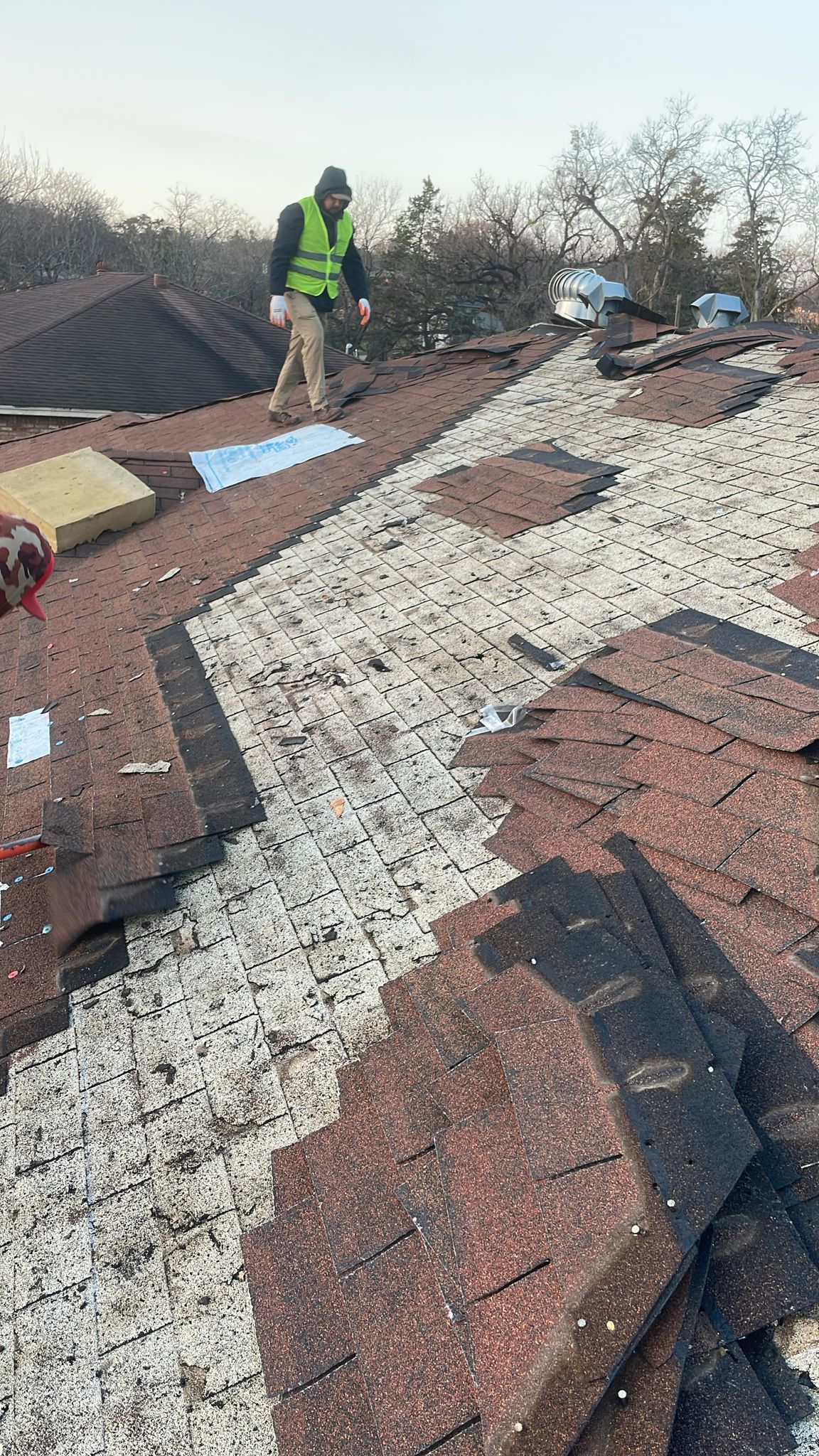 Person in safety vest inspects a damaged roof with scattered shingles and debris.