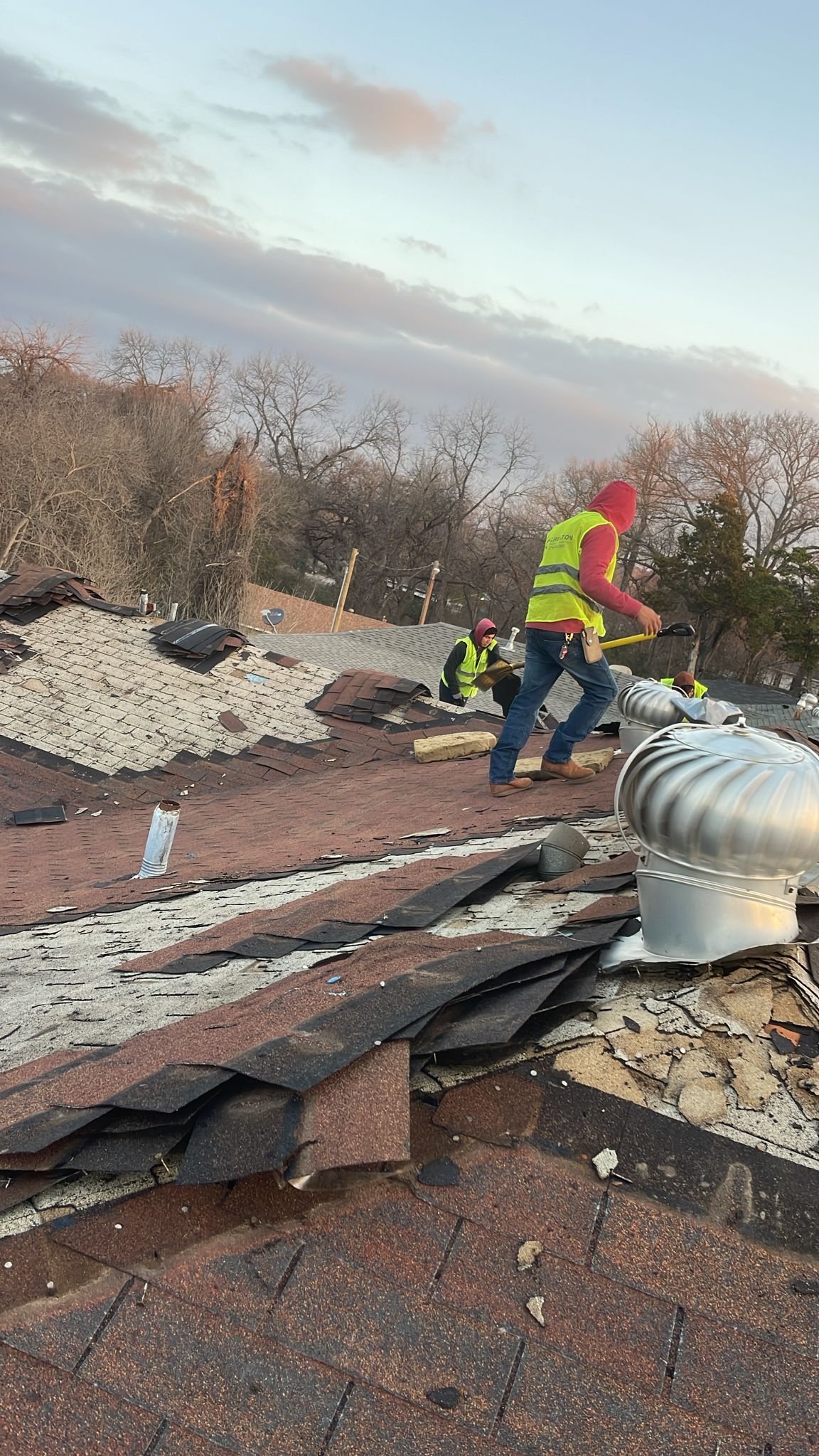 Two roofers in safety vests work on a damaged roof with a sunset in the background.