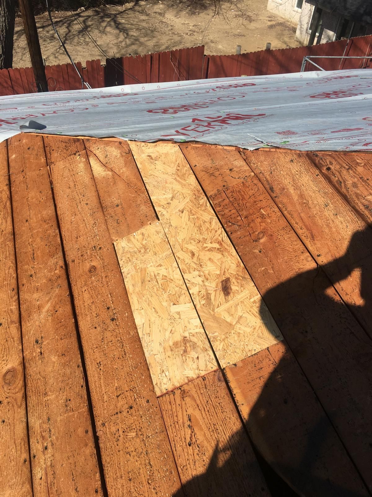 Wooden roof with a square patch of OSB; roofing paper and a fence in the background.