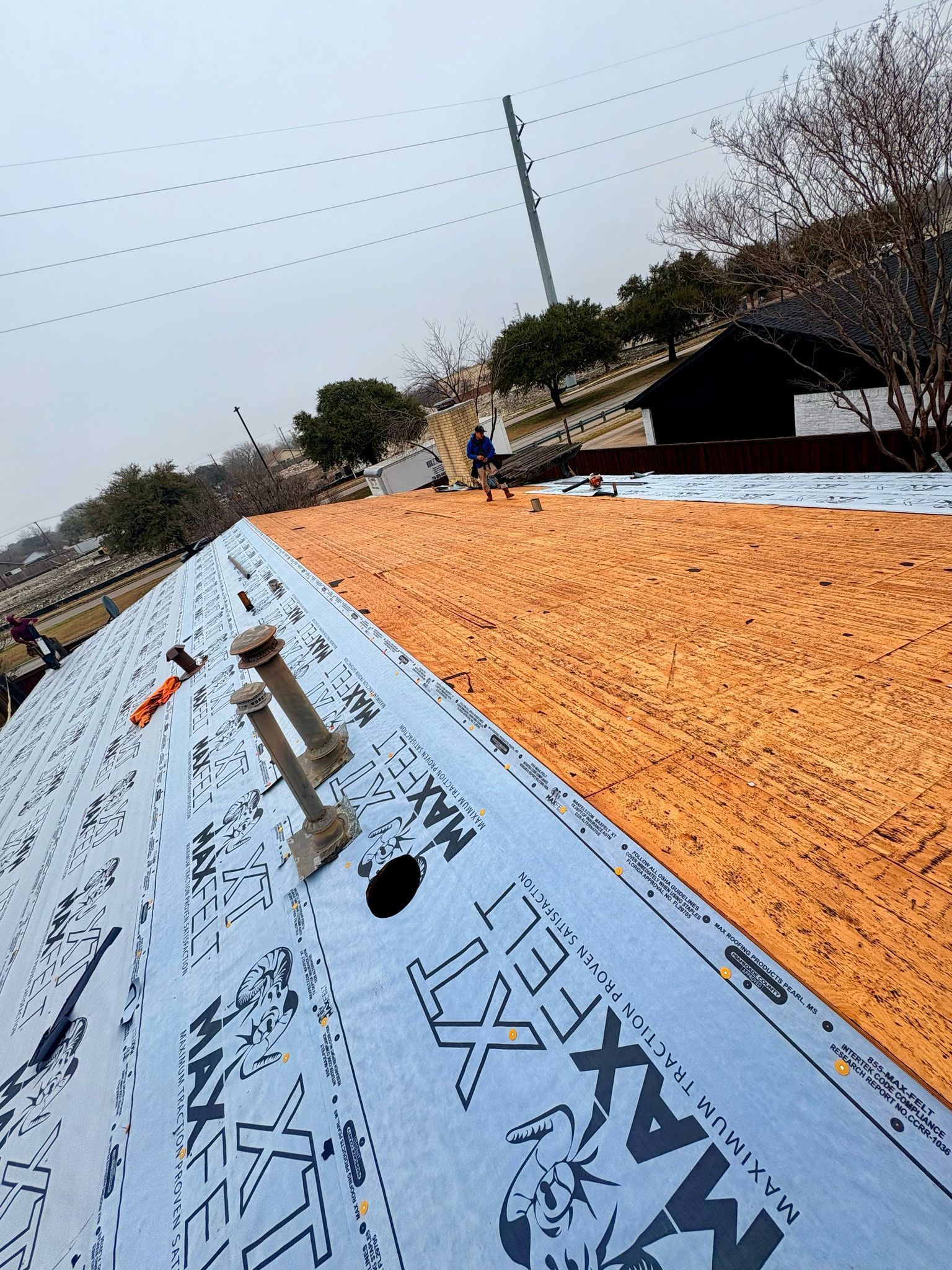 Rooftop with orange underlayment, blue roofing material, and construction worker in the background. Cloudy sky.
