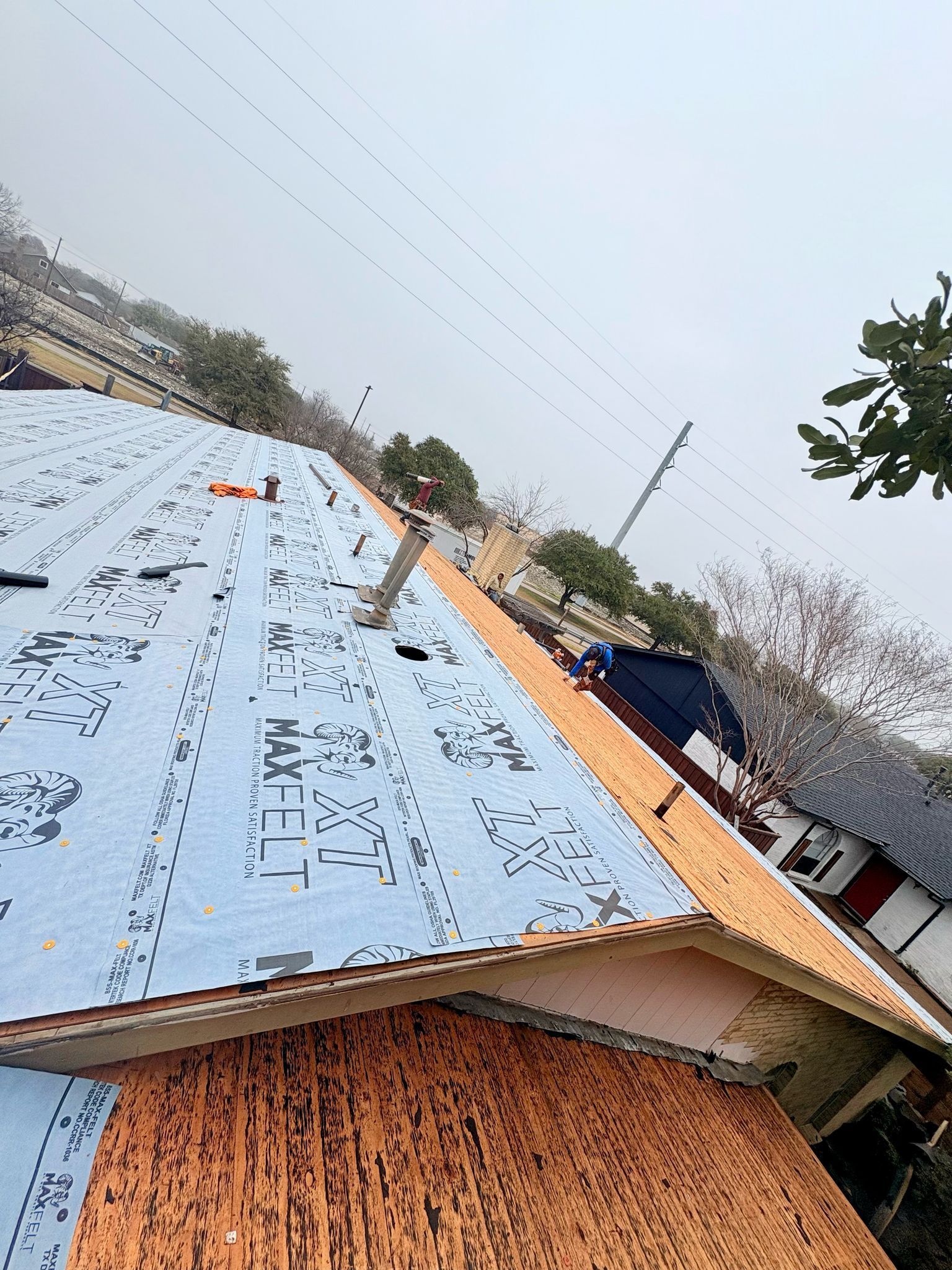 Roof being repaired, partially covered with blue underlayment and orange shingles. Chimney visible against a cloudy sky.