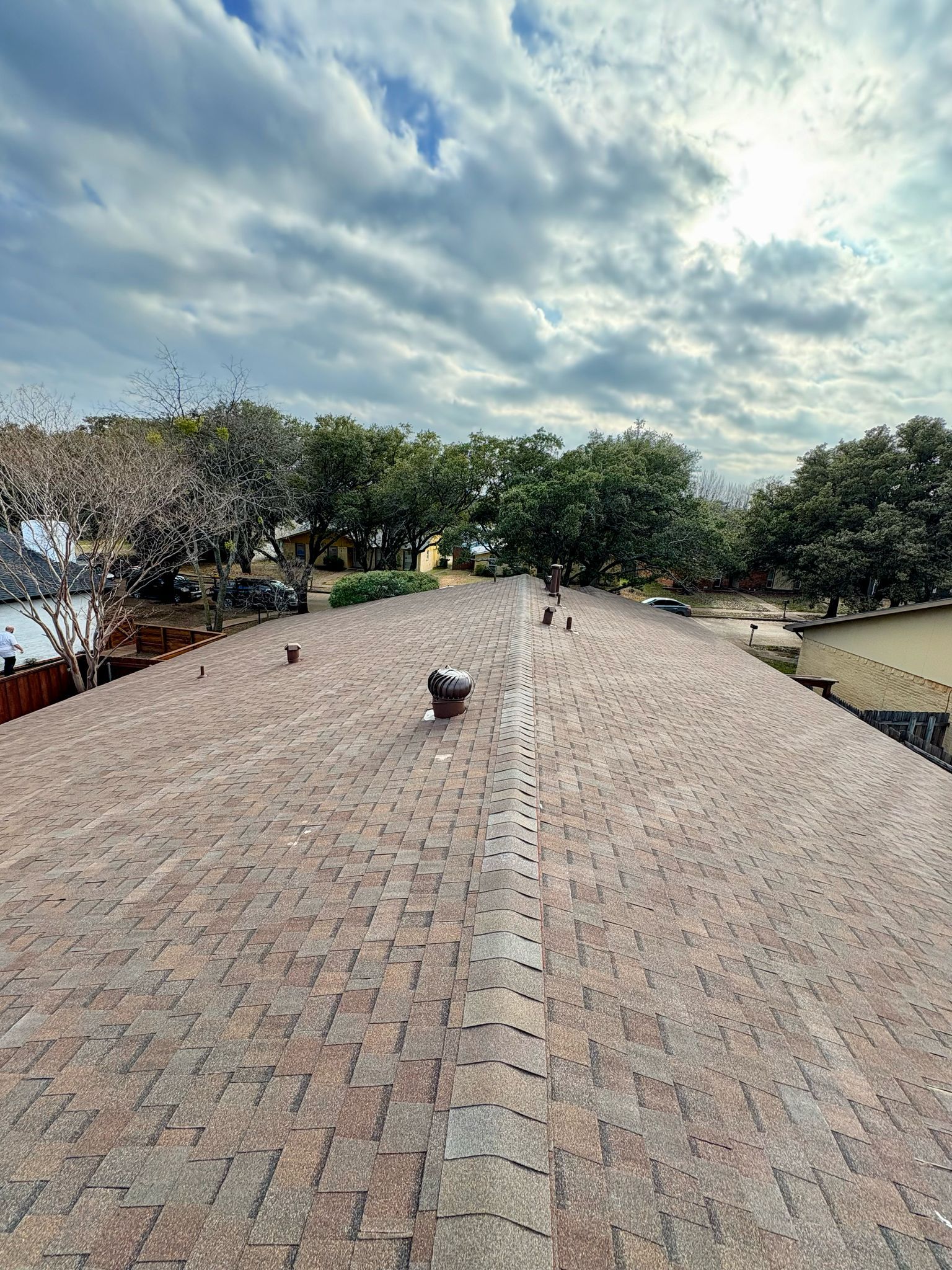 Brown shingled roof with a vent, trees, and a cloudy sky in the background.