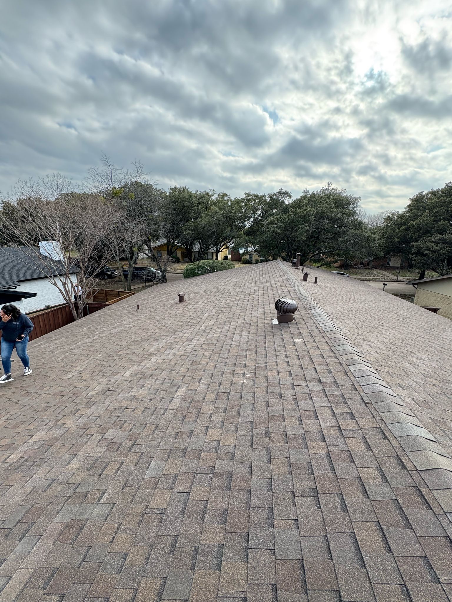 Person on a brown shingled roof under a cloudy sky. Trees and a house are in the background.