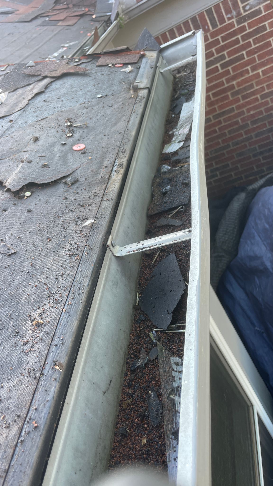 Gutter filled with debris and torn roof shingles next to a brick wall and window.