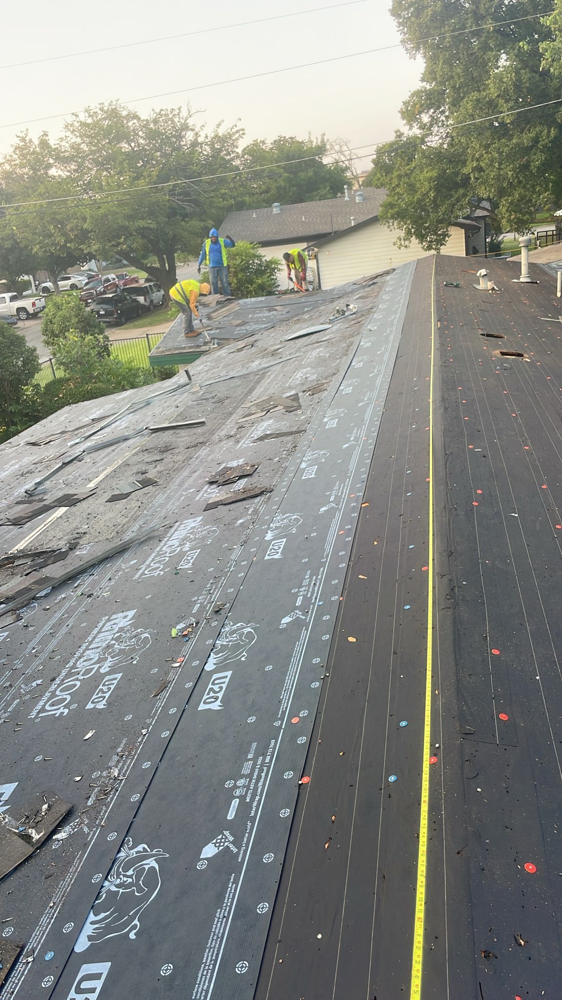 Workers on a roof installing shingles, asphalt surface, green trees in background, bright sunlight.