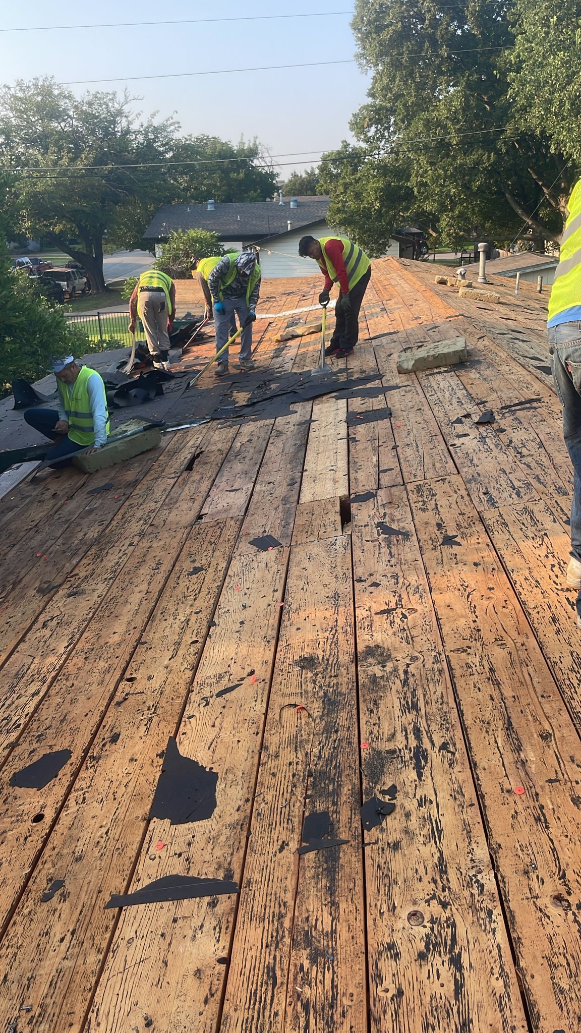 Construction workers on a roof, removing old decking.  They wear safety vests and are surrounded by trees.