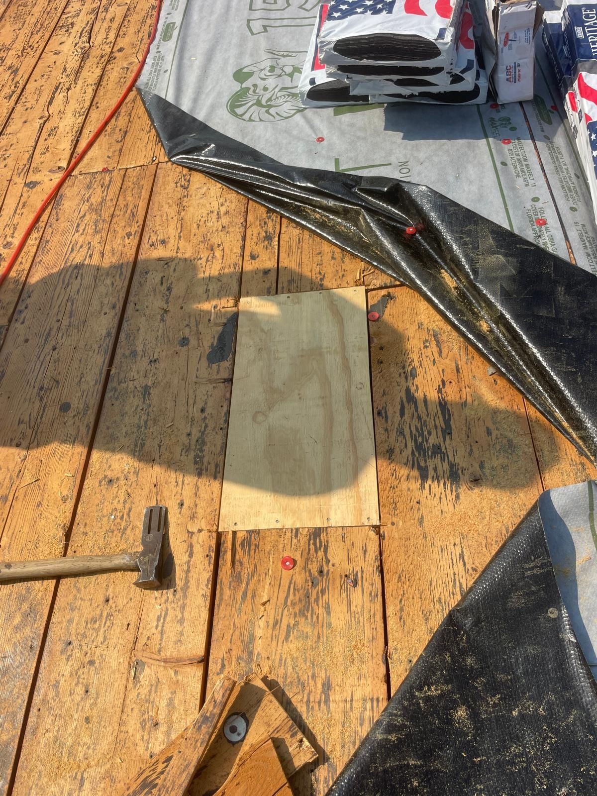 Wooden roof under construction; a piece of plywood sits on the roof, next to a hammer, and black roofing material.